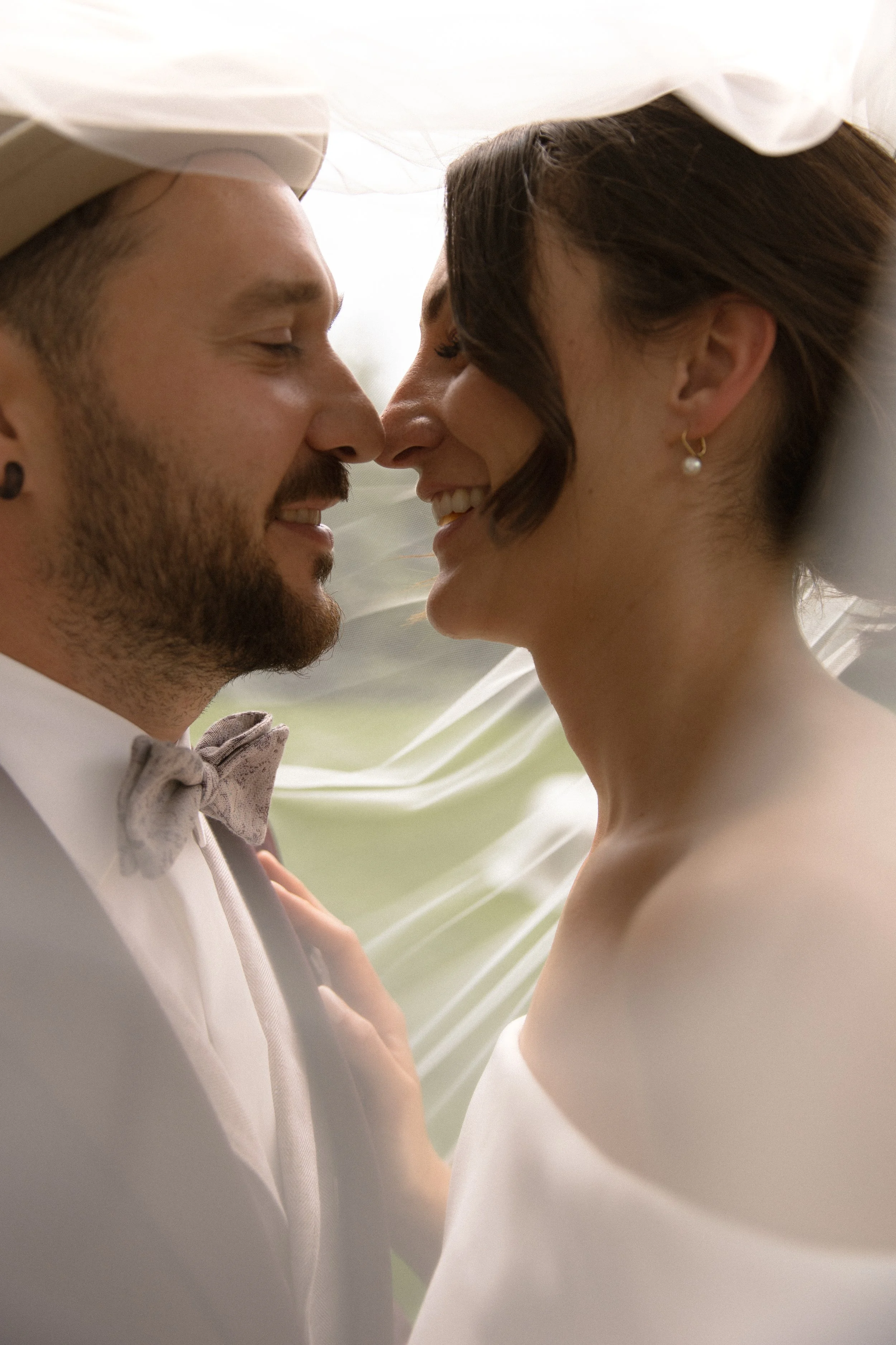 Ein verliebtes Paar bei ihrer Hochzeitszeremonie, das sich mit ihren Nasen berührt. Der Mann trägt einen weißen Anzug und Fliege, die Frau trägt ein weißes Kleid und Perlenohrhänger.