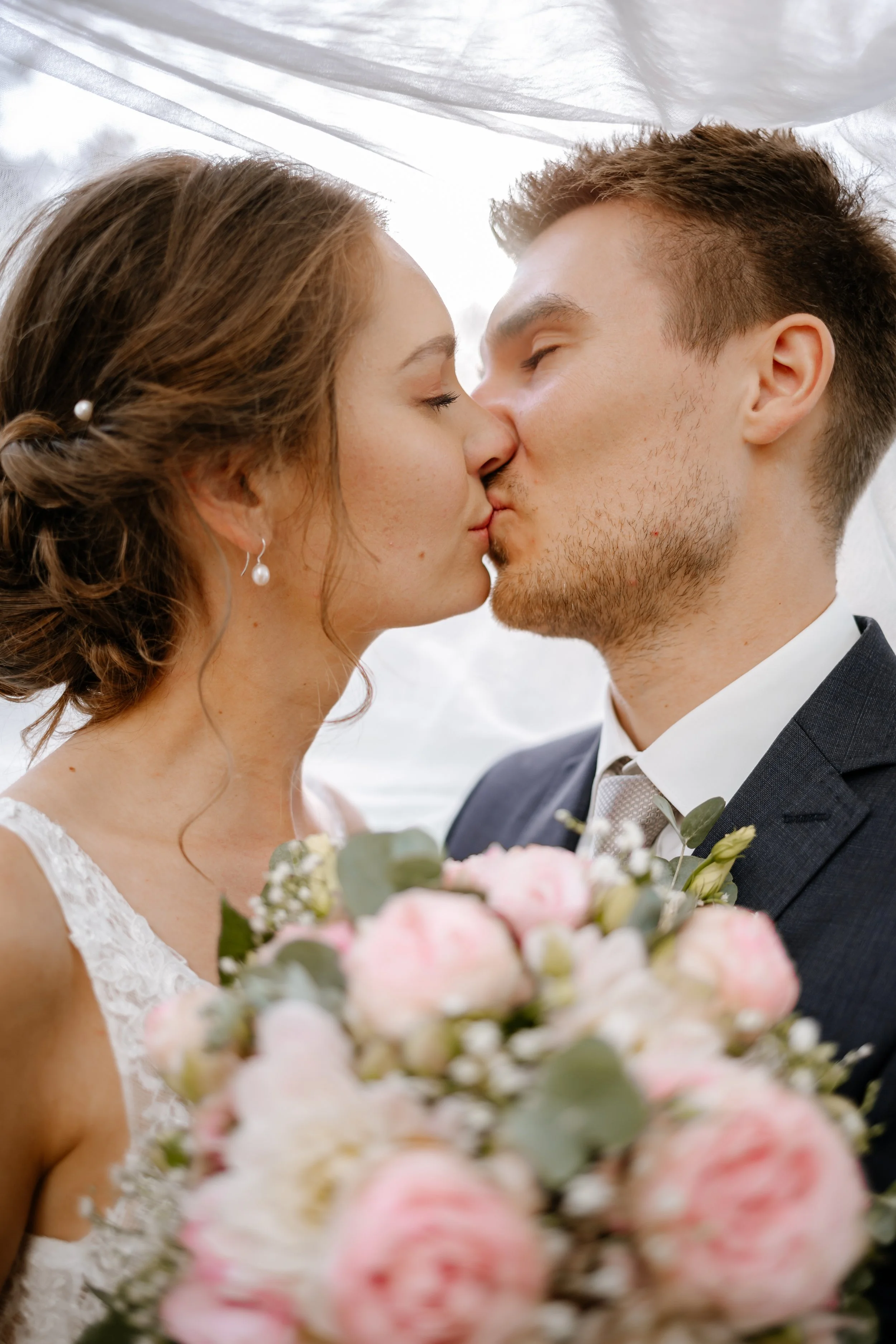 Ein Brautpaar küsst sich bei ihrer Hochzeit, mit einer Brautstrauß aus rosa und weißen Blumen und einer silbernen Haarspange im Haar der Braut.