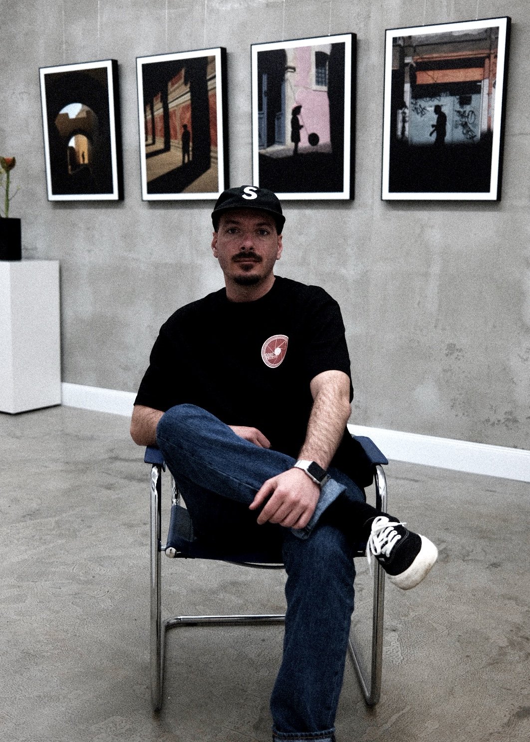 Ein junger Mann sitzt in einem Kunstmuseum vor einer Wand mit fünf Kunstfotografien. Er trägt eine schwarze Cap, ein schwarzes T-Shirt, Jeans und Turnschuhe, sitzt mit verschränkten Beinen und schaut in die Kamera.