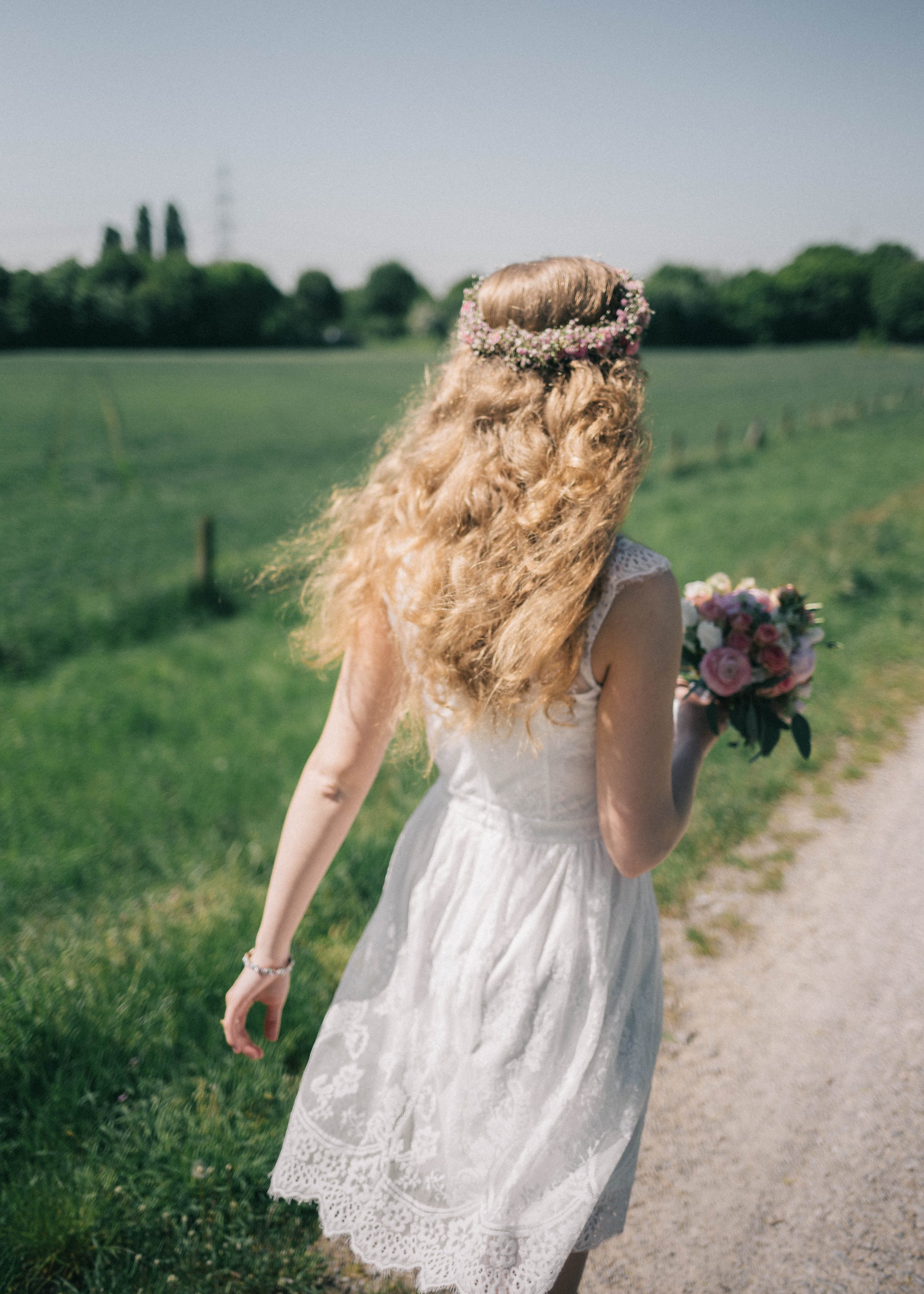 Junge Frau in weißem Kleid mit Blumenkranz im Haar, läuft auf Feldweg mit Blumenstrauß in der Hand, Blick nach hinten, sonniger Tag.