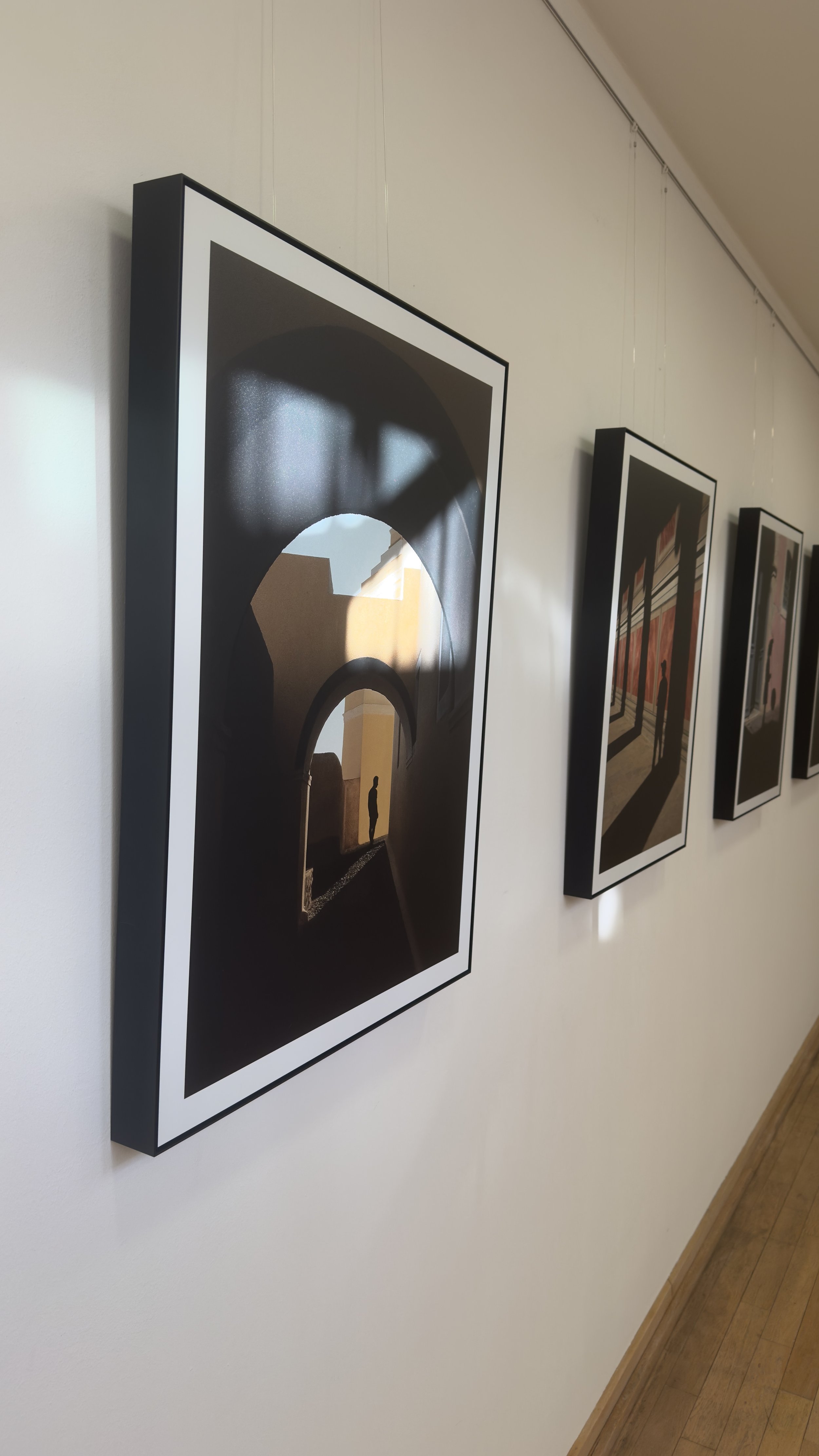 Fotografie einer Galeriewand mit mehreren Gemälden in schwarzen Rahmen, eines im Fokus zeigt einen dunklen Tunnel mit einem Menschen und architektonischen Elementen im Hintergrund.