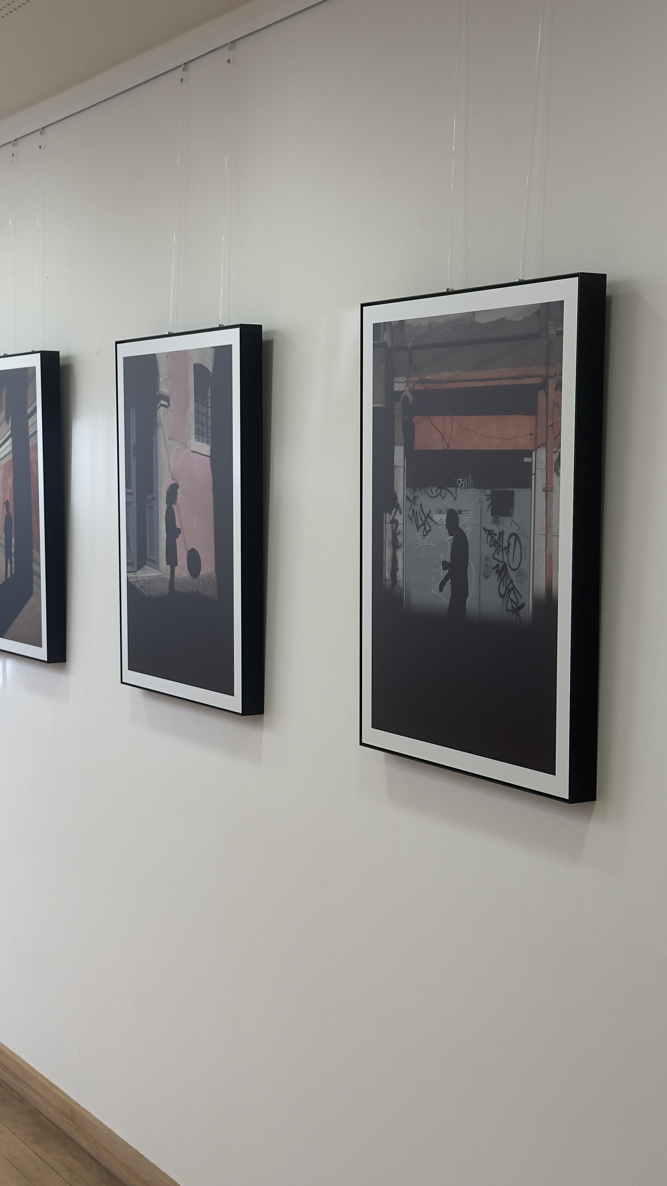 Drei gerahmte Schwarz-Weiß-Fotografien an einer weißen Wand in einer Galerie, die Silhouetten von Menschen in urbaner Umgebung zeigen.