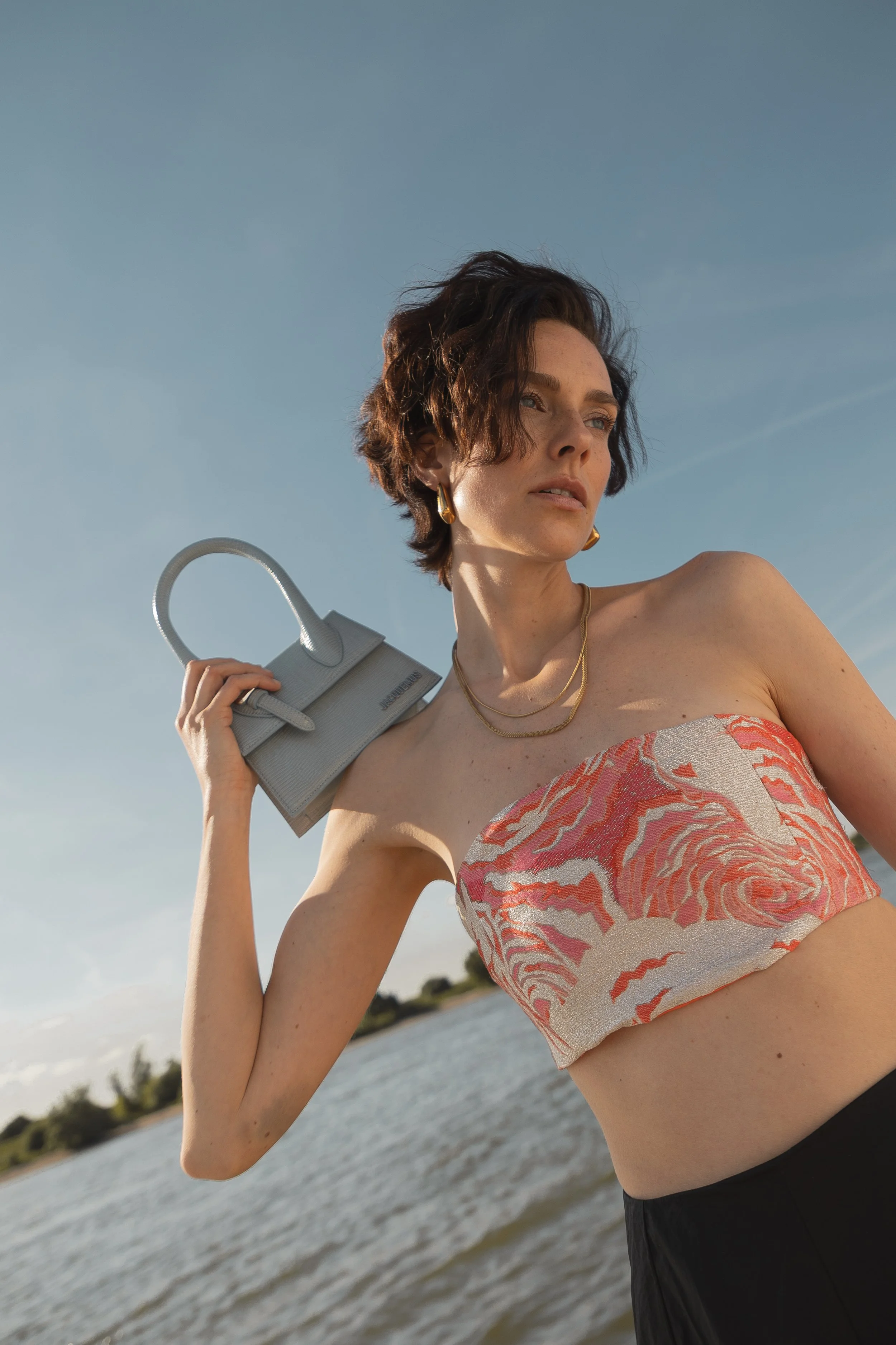 Frau am Wasser mit kurzer, welliger brauner Haarfrisur, trägt ein rotes und weißen Bandeau-Top, schwarze Hosen, goldene Ohrringe und Halsketten, hält eine graue Handtasche über die Schulter, bei Sonnenuntergang oder am späten Nachmittag.