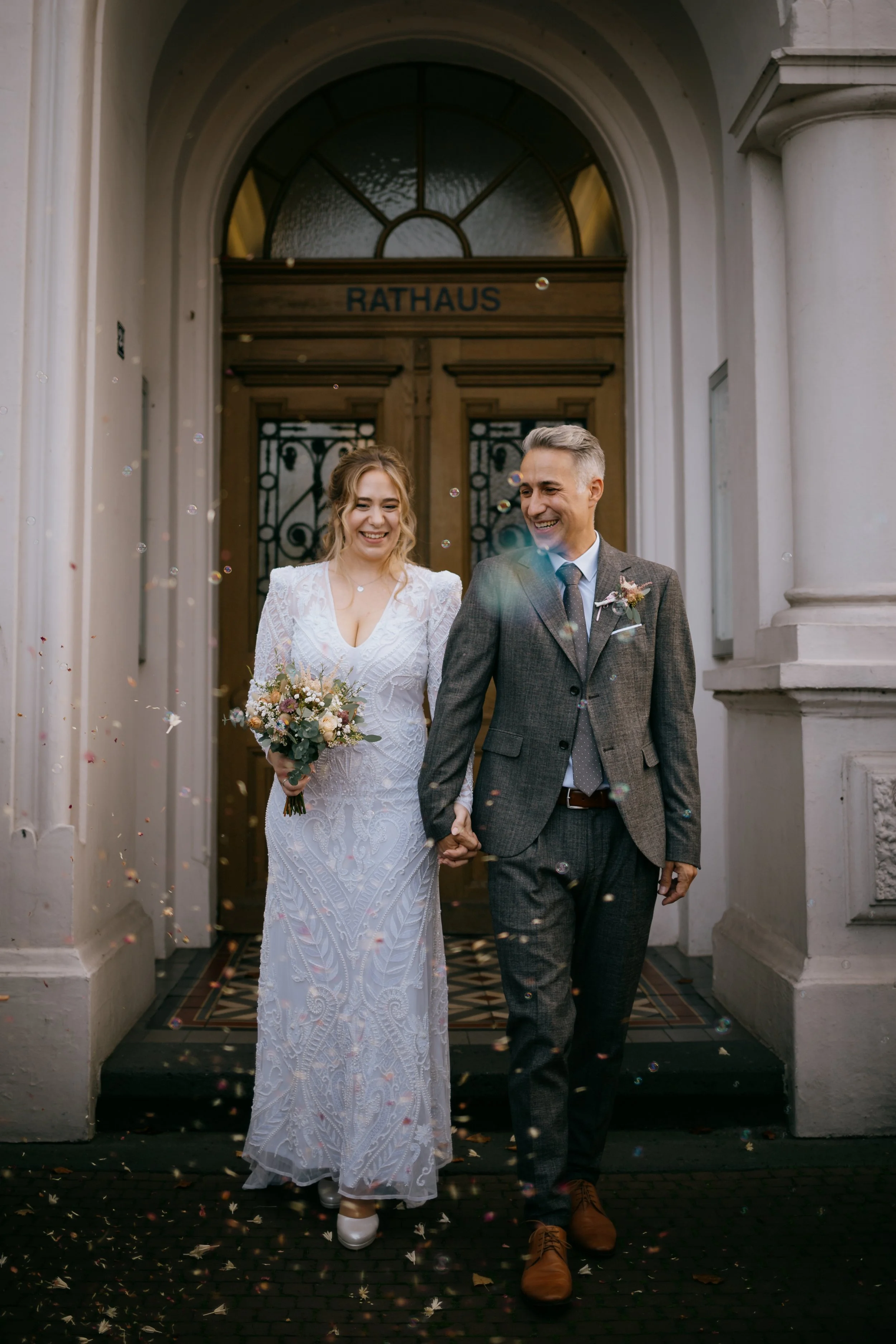 Ein frisch verheiratetes Paar geht an der Tür eines Rathauses vorbei, lächelnd und Händchen haltend, während Konfetti um sie herum fliegt.