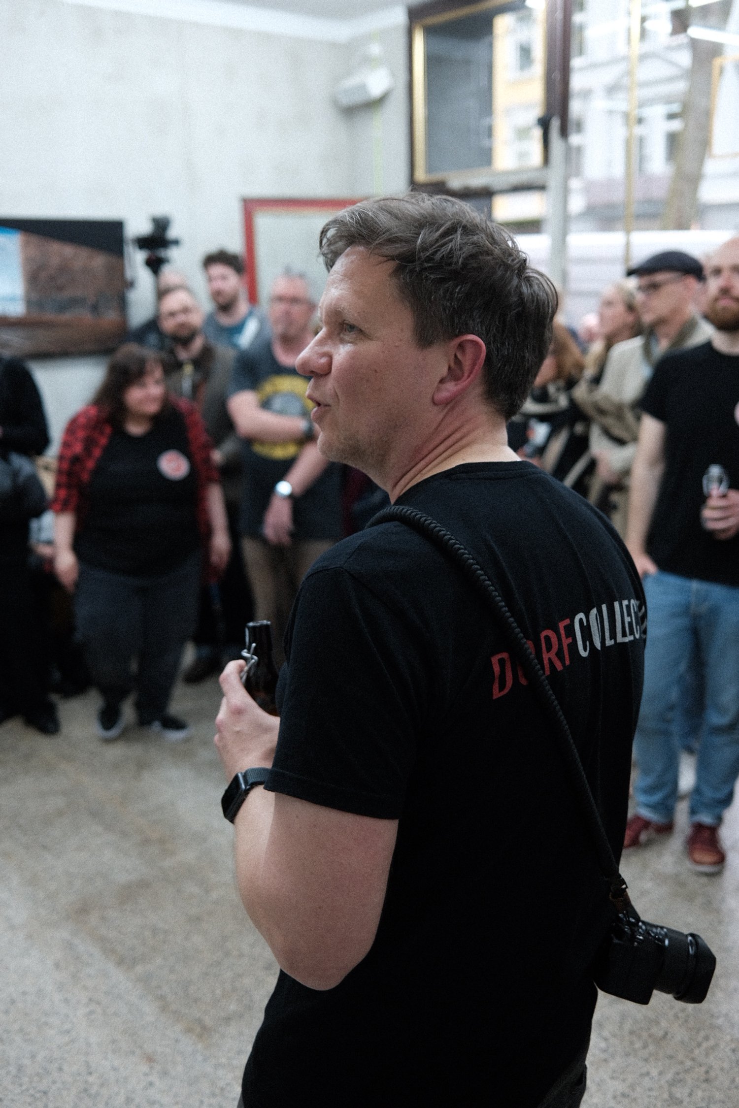 Ein Mann in schwarzem T-Shirt mit Kamera um den Hals, umgeben von einer Gruppe von Menschen in einem Raum.