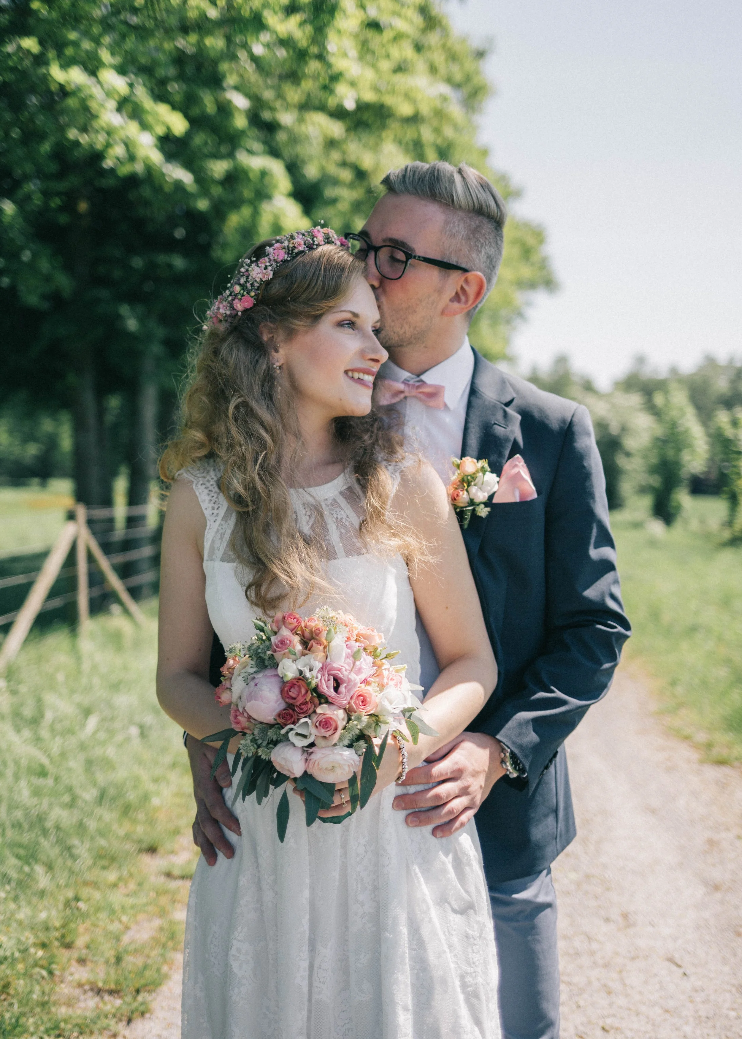 Ein Brautpaar steht im Freien auf einer Wiese, die Braut trägt ein weißes Hochzeitskleid mit Spitzen und hält einen Blumenstrauß, der Bräutigam trägt einen Anzug mit pinker Fliege und Anstecker, er küsst die Braut auf die Stirn, hinter ihnen sind grü