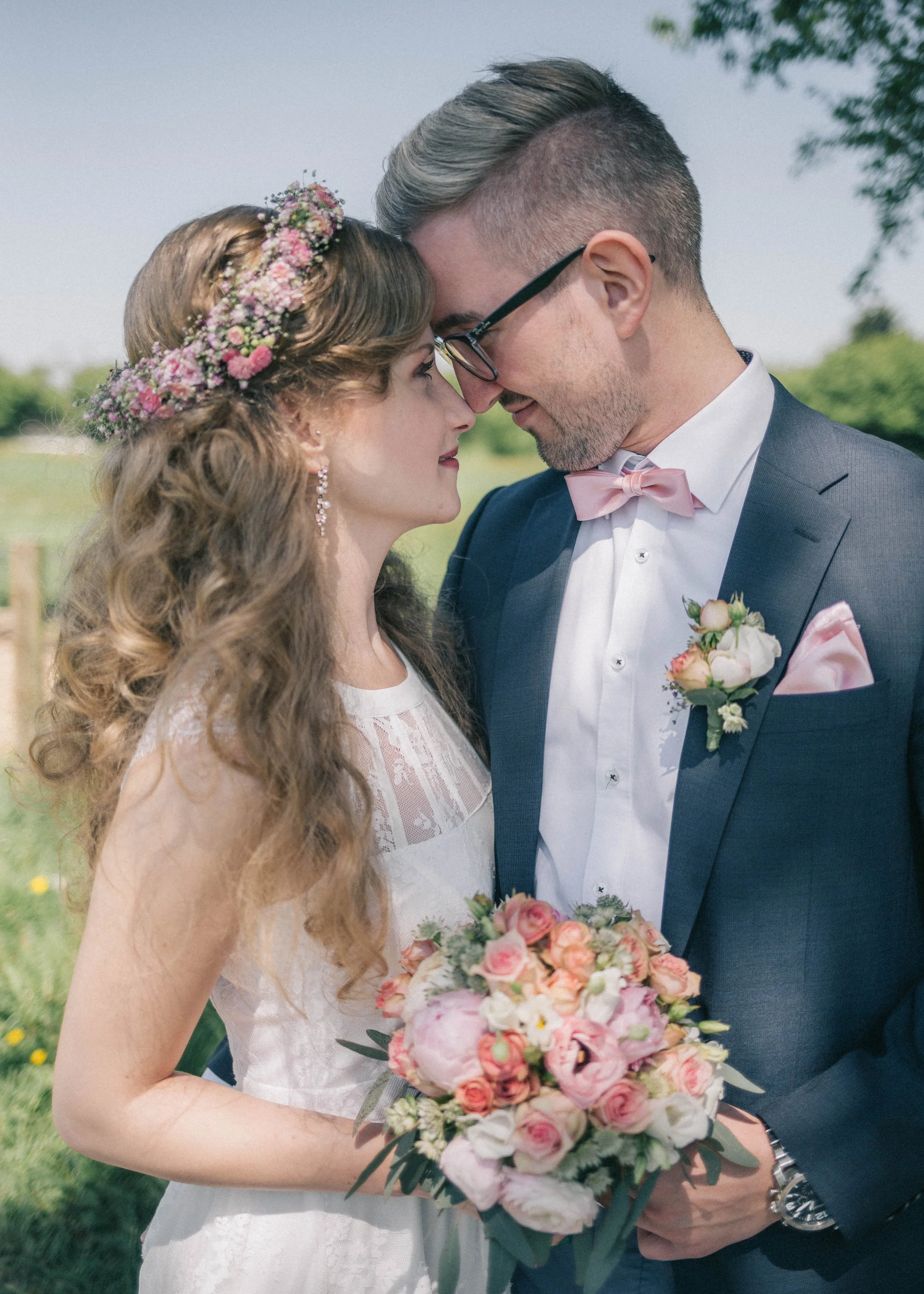 Ein frisch verheiratetes Paar steht im Freien, die Braut trägt einen Blumenkranz im Haar und hält einen Blumenstrauß, während das Paar sich mit Schultern berührt und sich liebevoll ansieht.