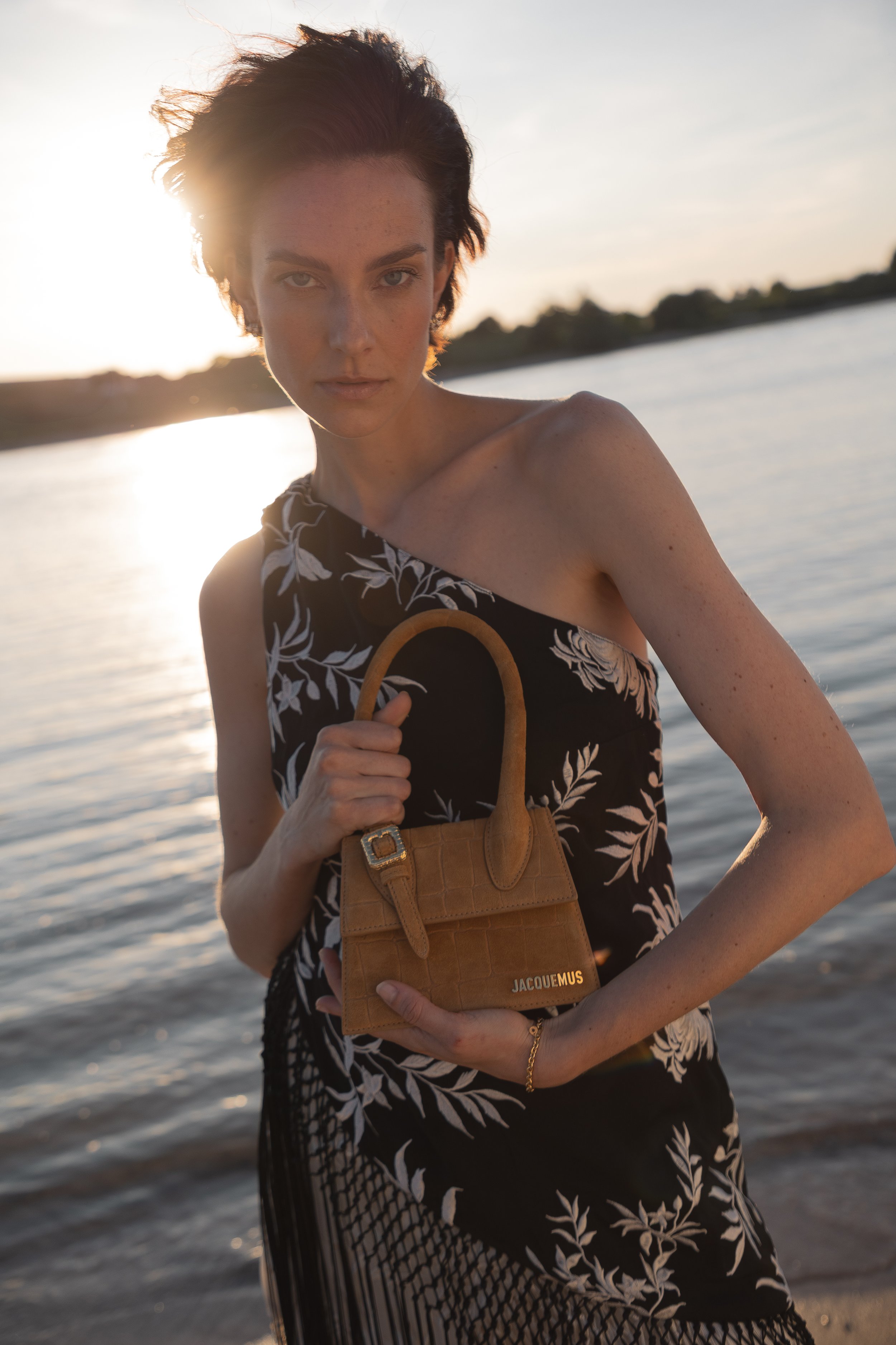 Junge Frau steht am Wasser bei Sonnenuntergang, hält eine braune Handtasche von Jacquemus, trägt ein schwarzes Kleid mit weißen Mustern.