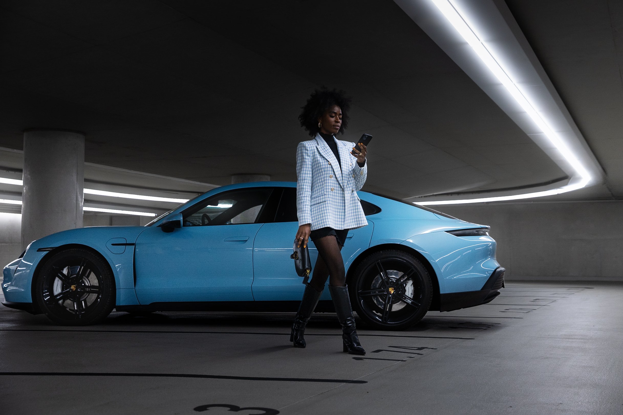 Frau in modischer Kleidung steht in einer Tiefgarage vor einem blauen Sportwagen und schaut auf ihr Smartphone.