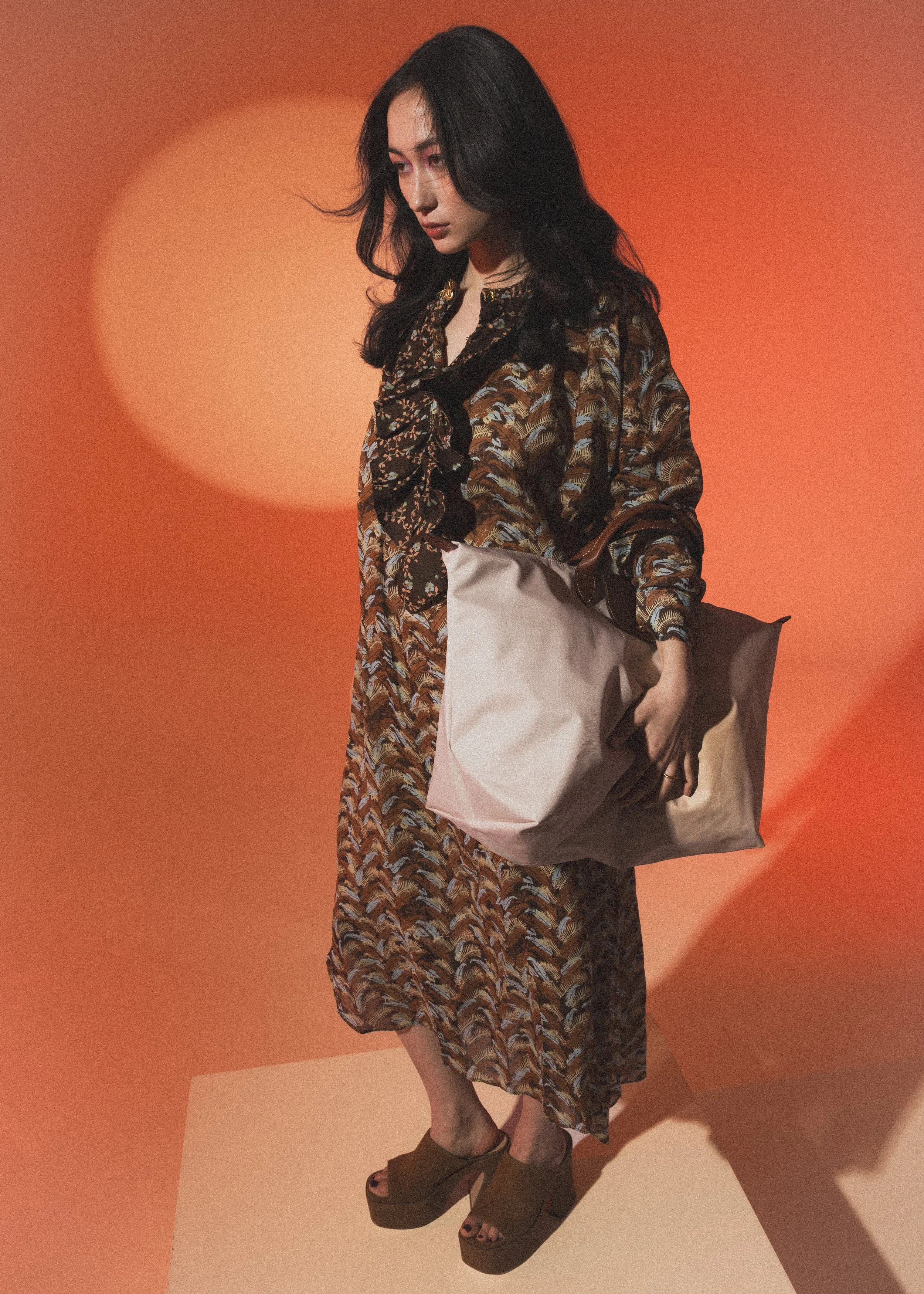 Junge Frau in einem langen, bunten Kleid, mit dunklen Haaren, trägt eine große Tasche und steht vor einem orangefarbenen Hintergrund.