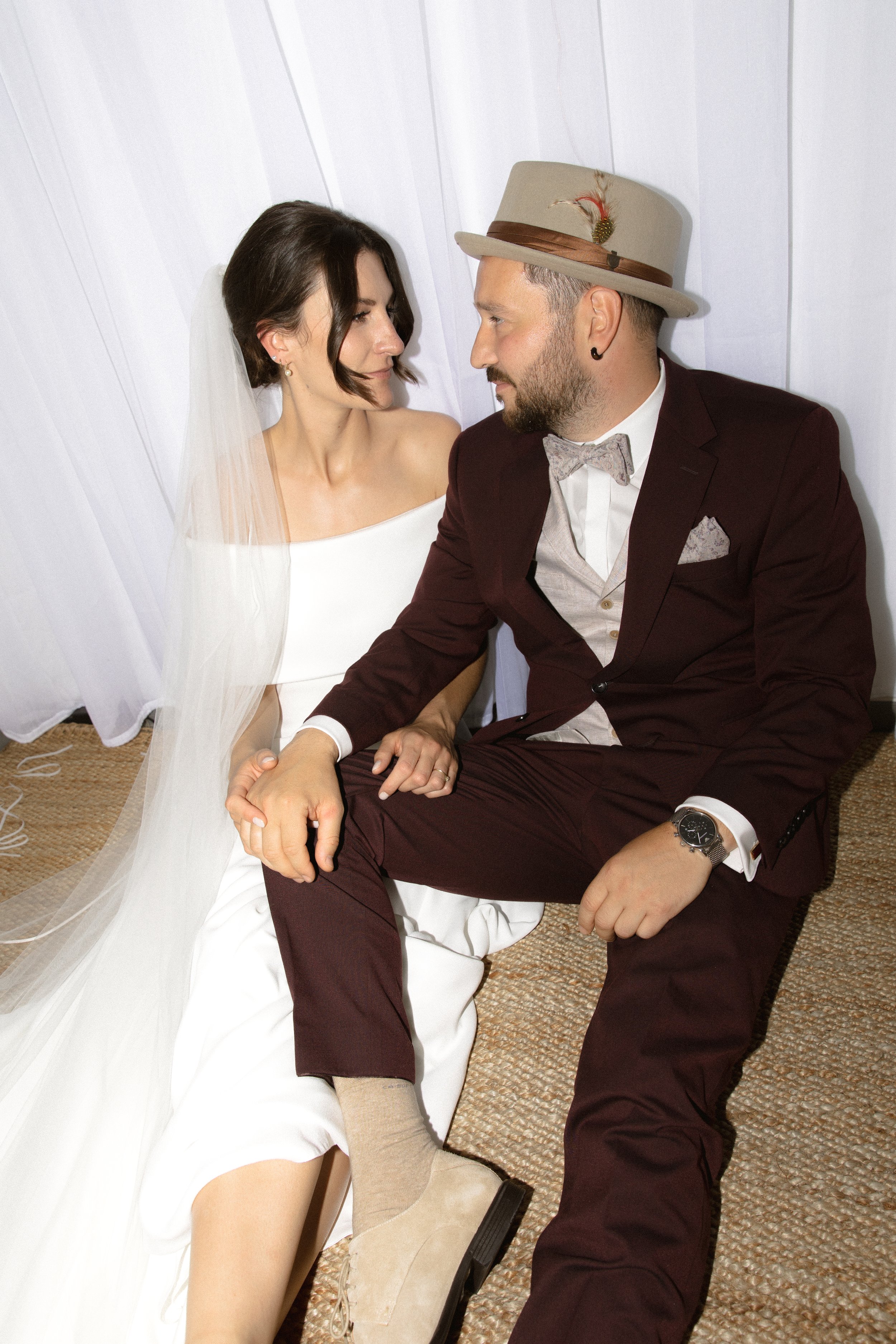Ein Brautpaar sitzt eng zusammengesunken auf dem Boden, die Braut trägt ein weißes Kleid mit Schleier, der Bräutigam trägt Anzug und einen Hut mit Feder, beide schauen sich liebevoll an.