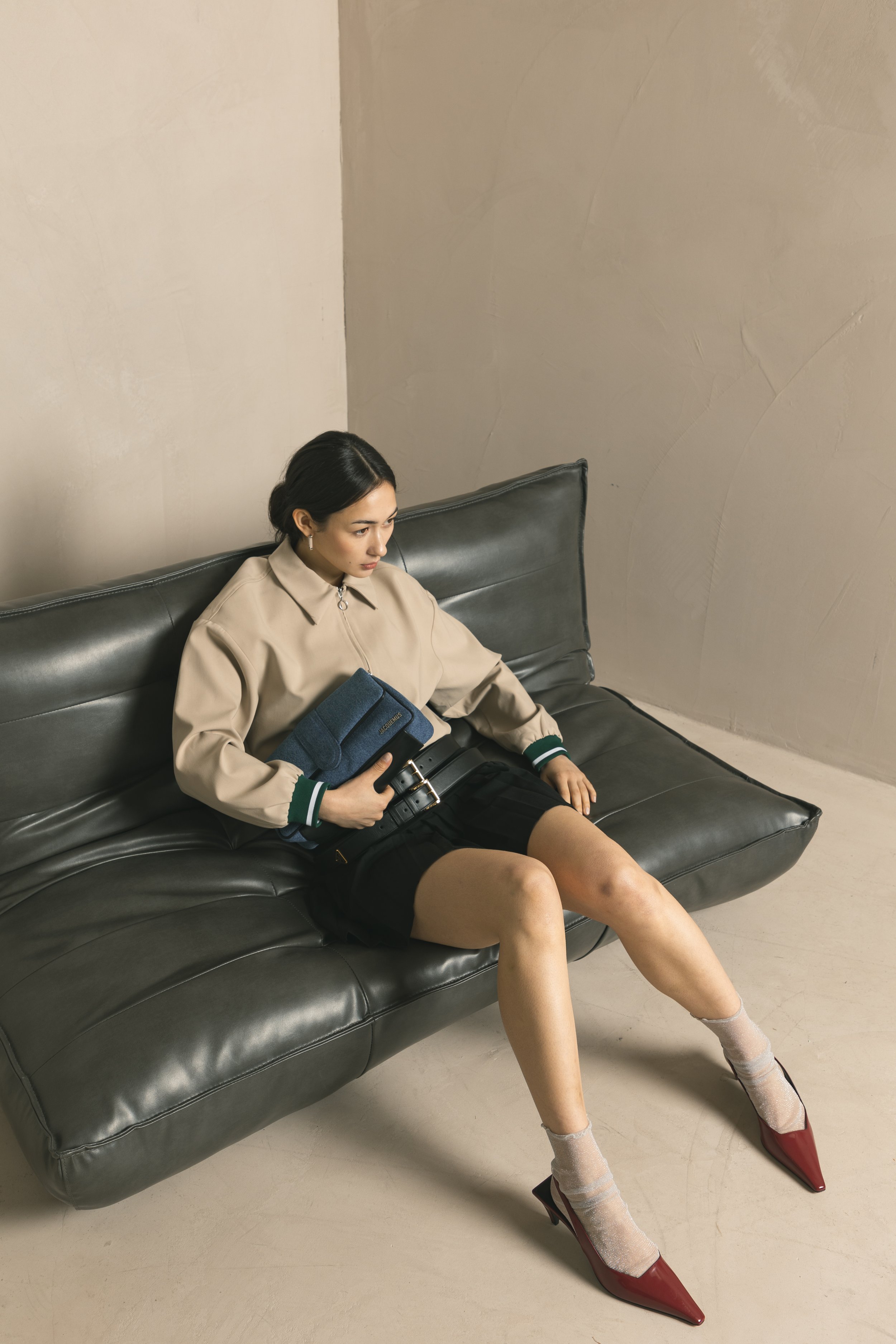 Junge Frau sitzt auf einem schwarzen Ledersofa, trägt beige Jacke, schwarze Shorts, weiße Socken und rote Pumps, hält eine blaue Tasche und schaut nachdenklich nach links, im Hintergrund eine graue Wand.