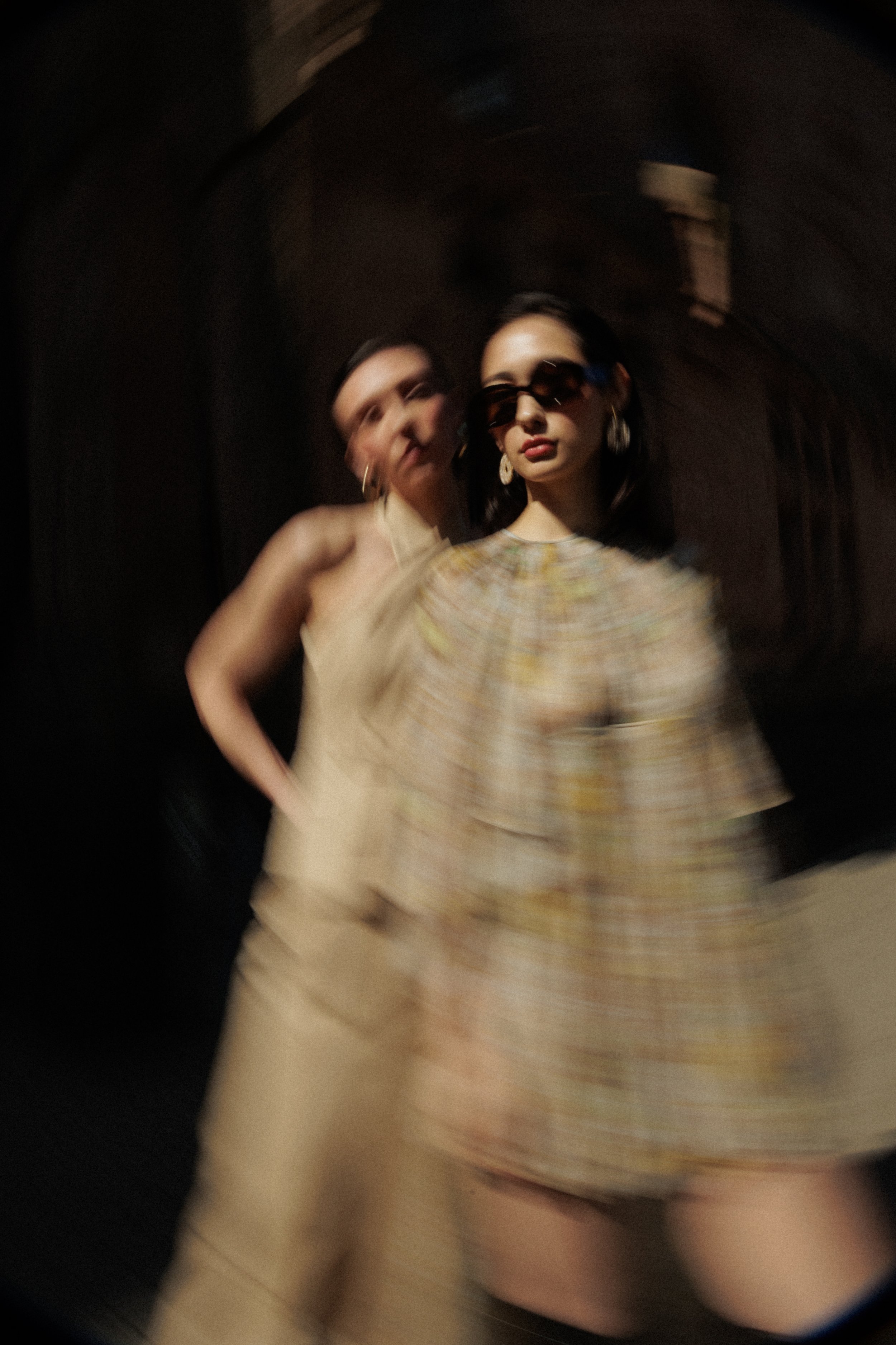 Zwei stilvoll gekleidete Frauen posieren elegant vor einem dunklen Hintergrund. Eine trägt eine beige Kleidung und die andere eine karierte Kleidung mit großen Sonnenbrillen. Die Fotos sind absichtlich unscharf und verschwommen.