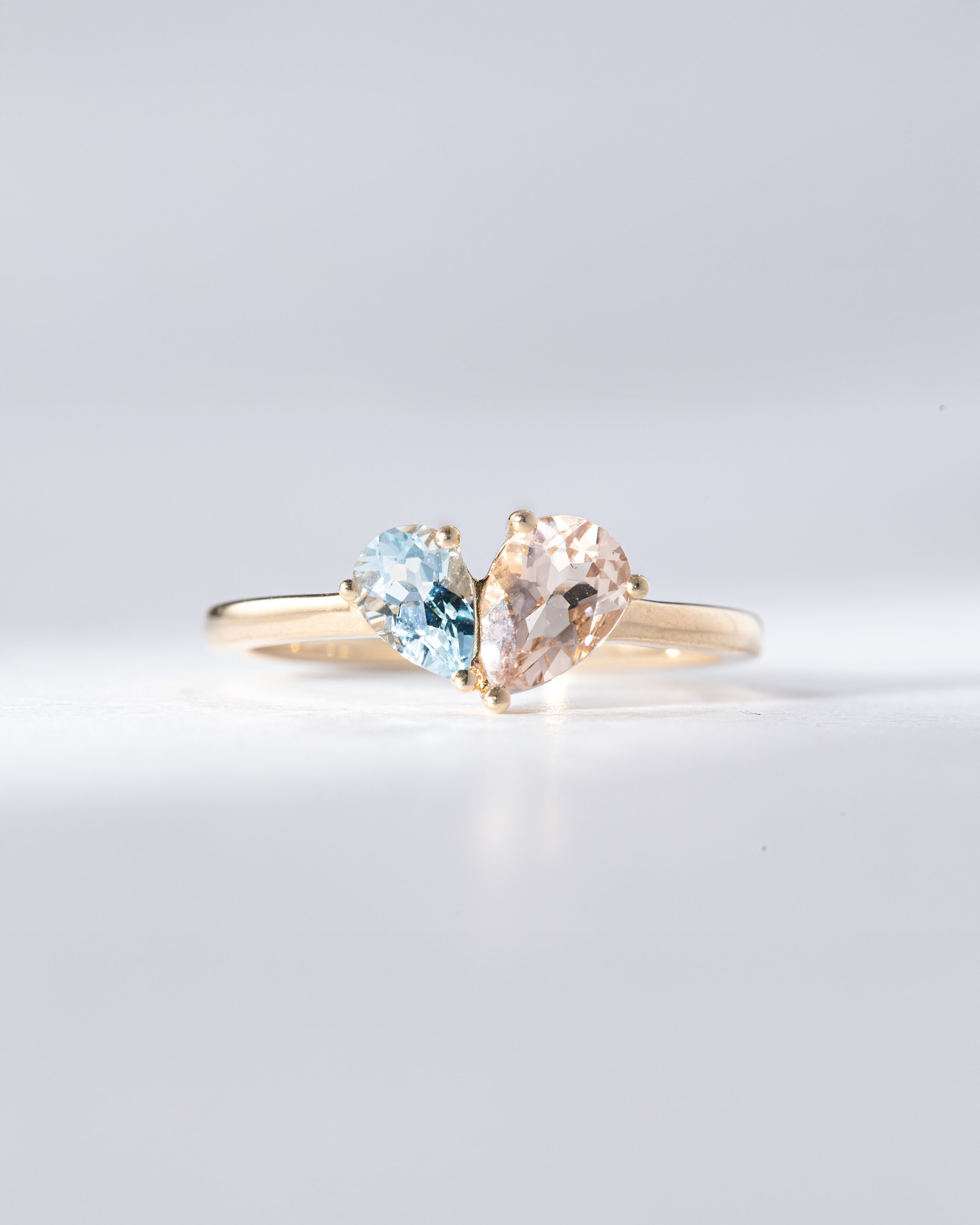 Goldring mit zwei herzförmigen, facettierten Edelsteinen, blau und rosafarben, auf weißem Hintergrund.