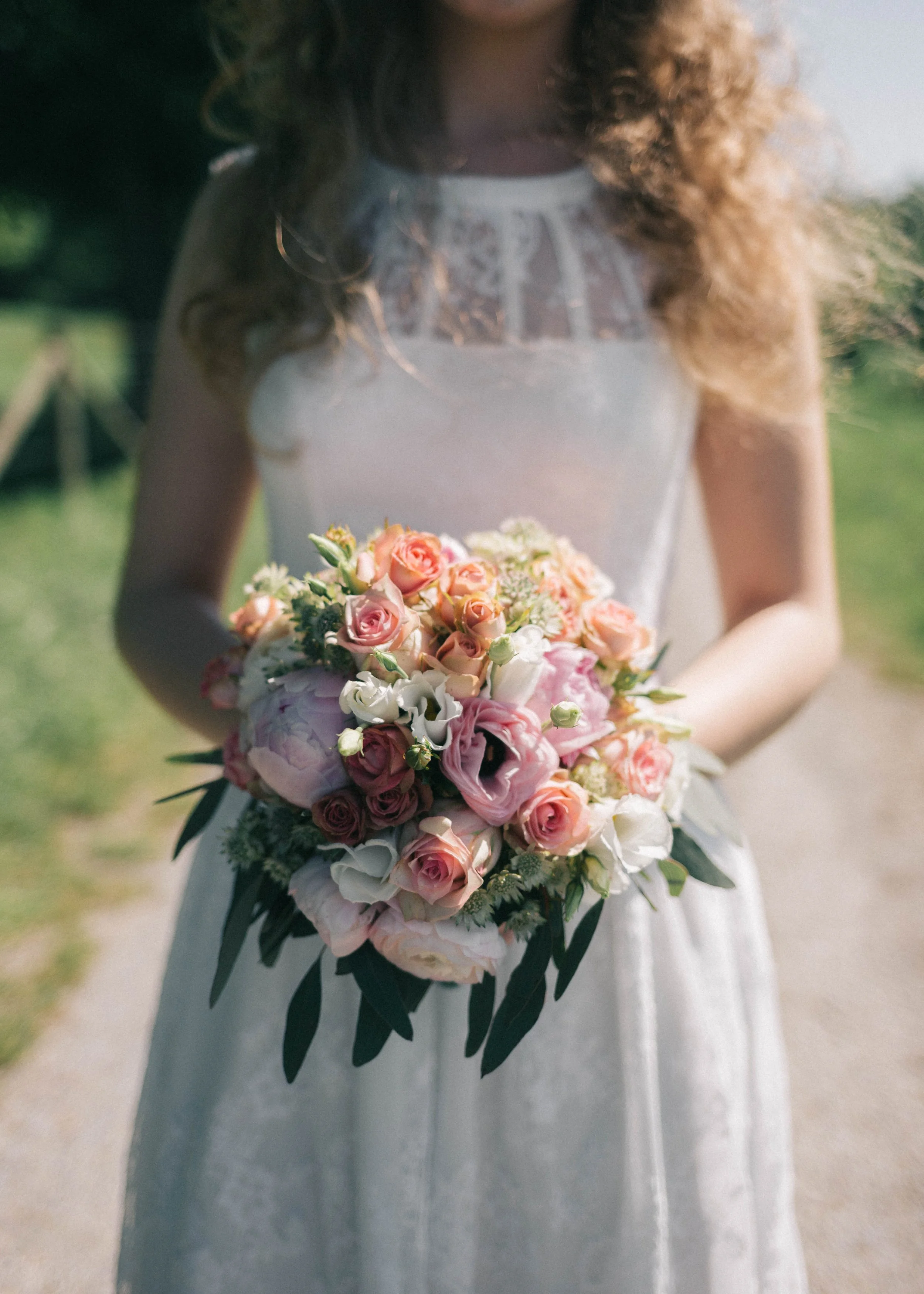 Frau in weißem Kleid, die einen Blumenstrauß aus rosa und weißen Blumen hält, draußen im Grünen.