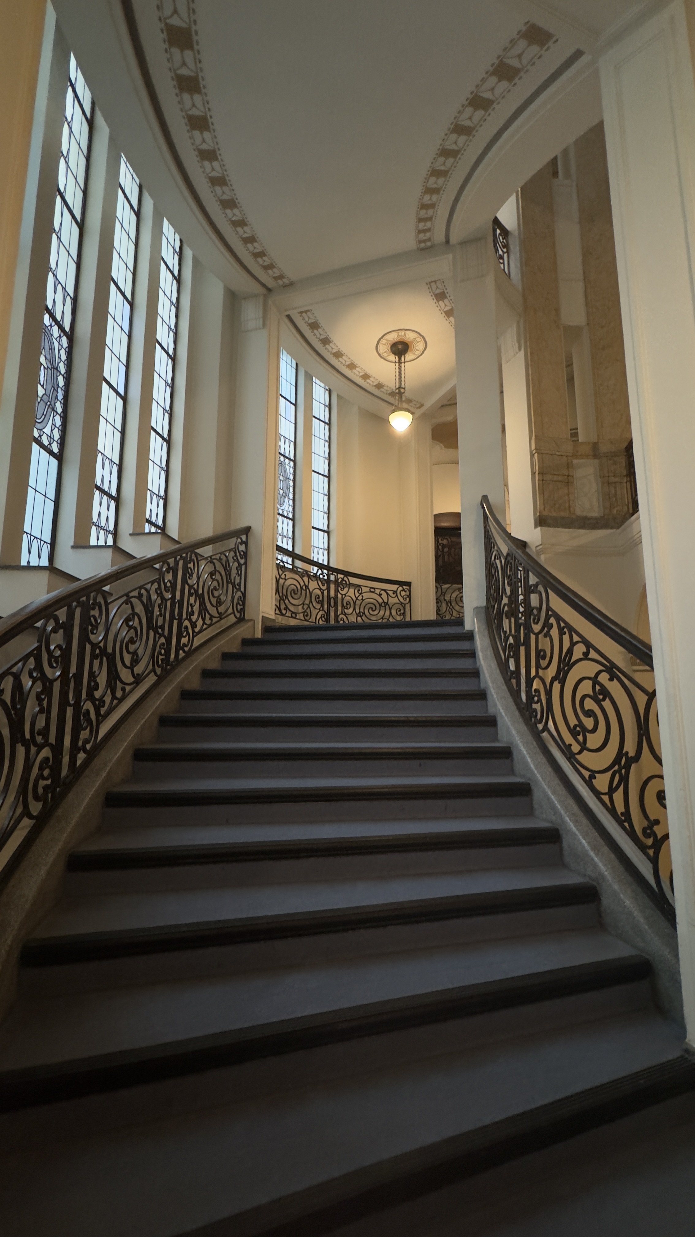 Innentreppenhaus mit geschwungenem Treppe, verzierte Schmiedereisenhandläufe, hohe Fenster mit Muster, Deckenleuchte, dekorative Deckenbänder.