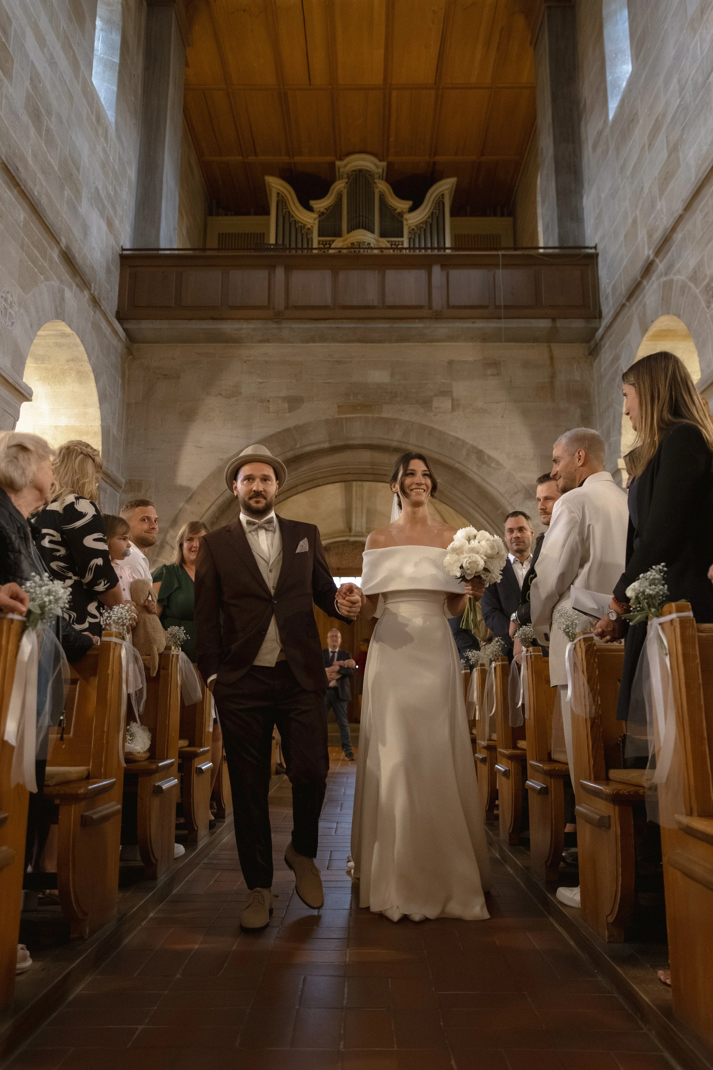 Ein Brautpaar läuft Hand in Hand durch eine Kirche, umgeben von Gästen. Der Altar im Hintergrund und die festliche Atmosphäre sind sichtbar.