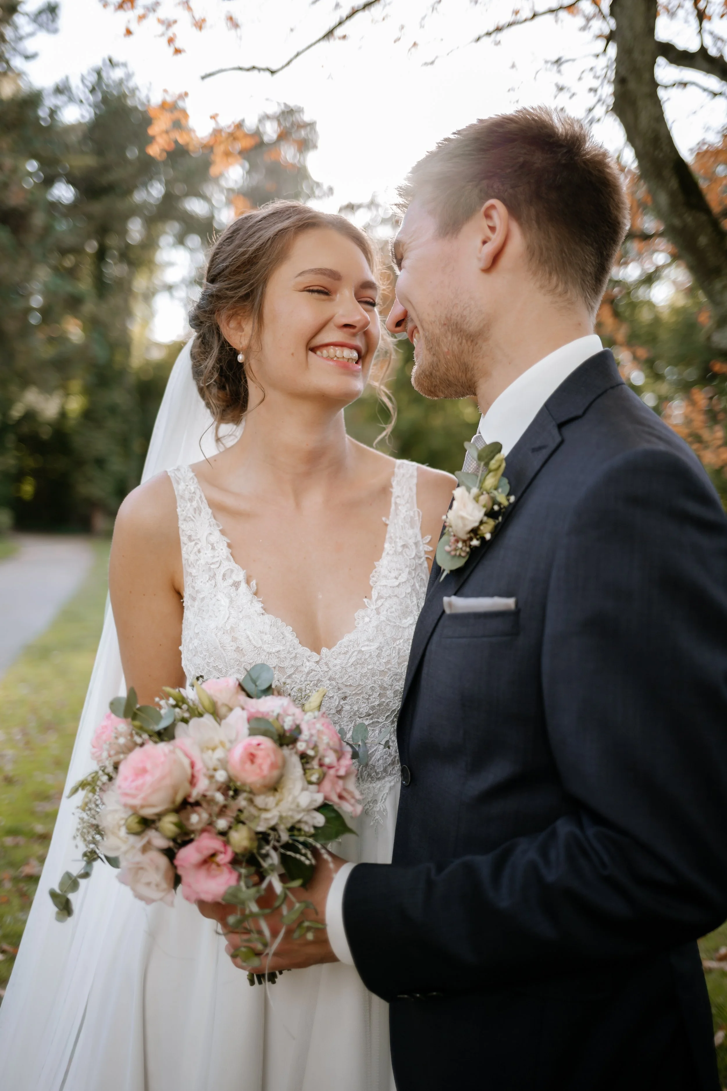 Ein glückliches Brautpaar steht im Park, lächelt sich verliebt an, die Braut trägt ein weißes Hochzeitskleid mit Spitze, hält einen Blumenstrauß mit rosa und weißen Blumen, die Umgebung ist herbstlich mit bunten Blättern an den Bäumen.
