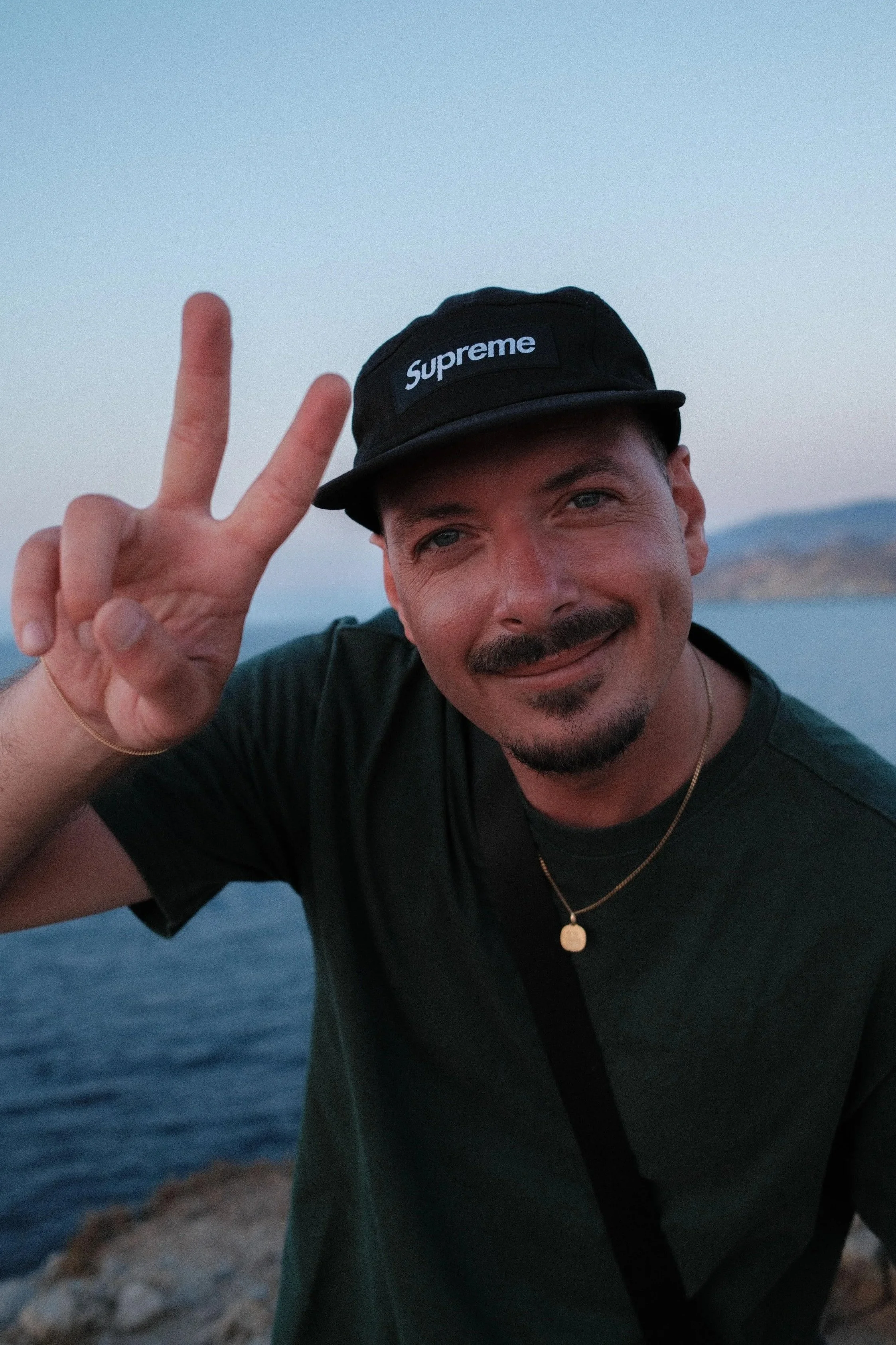 Ein lachender Mann mit dunklem Haarschopf, Bart und Schnurrbart trägt eine schwarze Cap mit "Supreme"-Logo, macht ein Peace-Zeichen, steht vor Meer und Bergen im Sonnenuntergang.