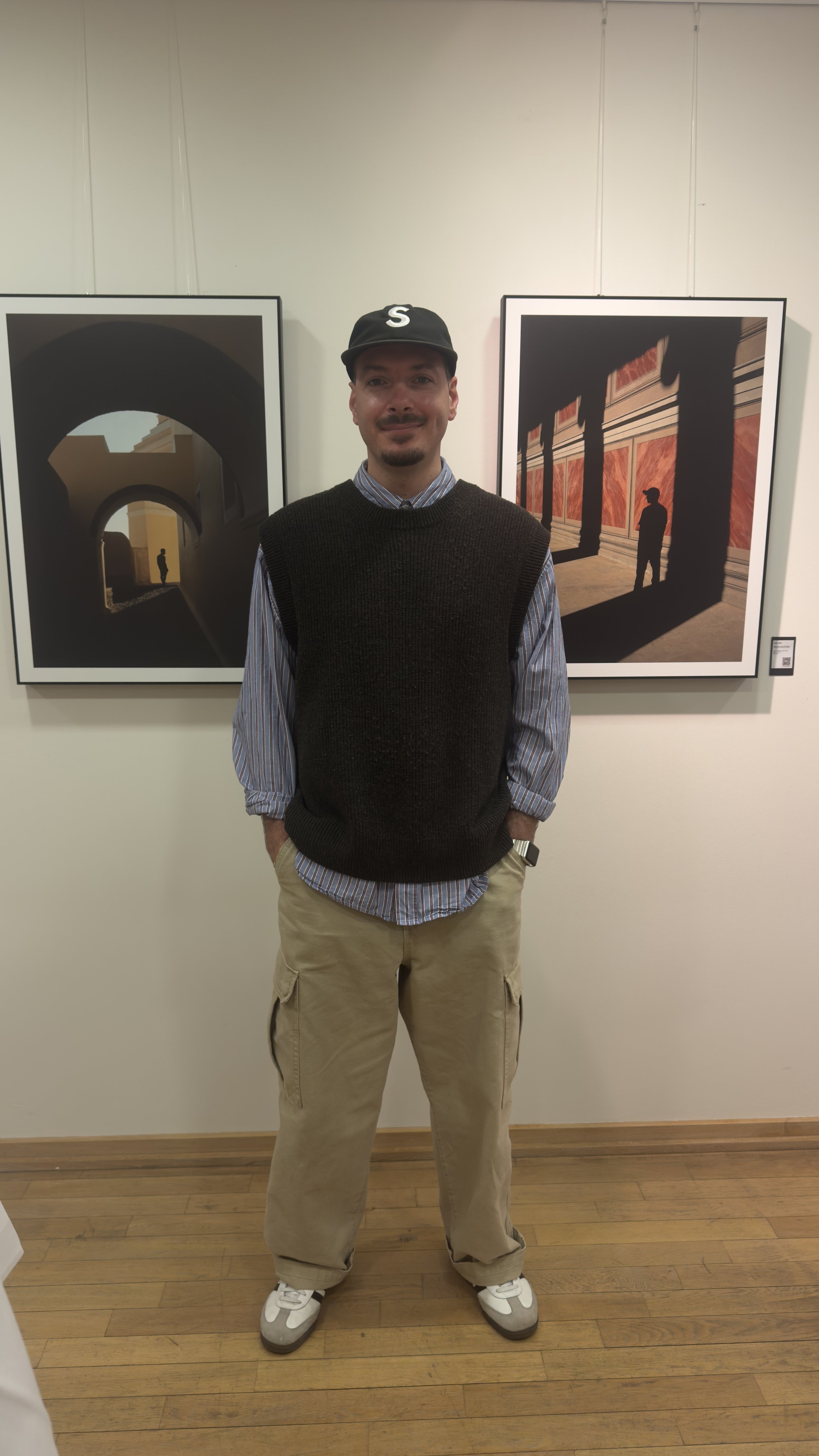 Ein Mann trägt einen schwarzen Cap mit einem weißen S, ein gestreiftes Hemd, eine schwarze Weste und beige Cargo-Hosen, steht vor zwei Kunstwerken in einem Museum.