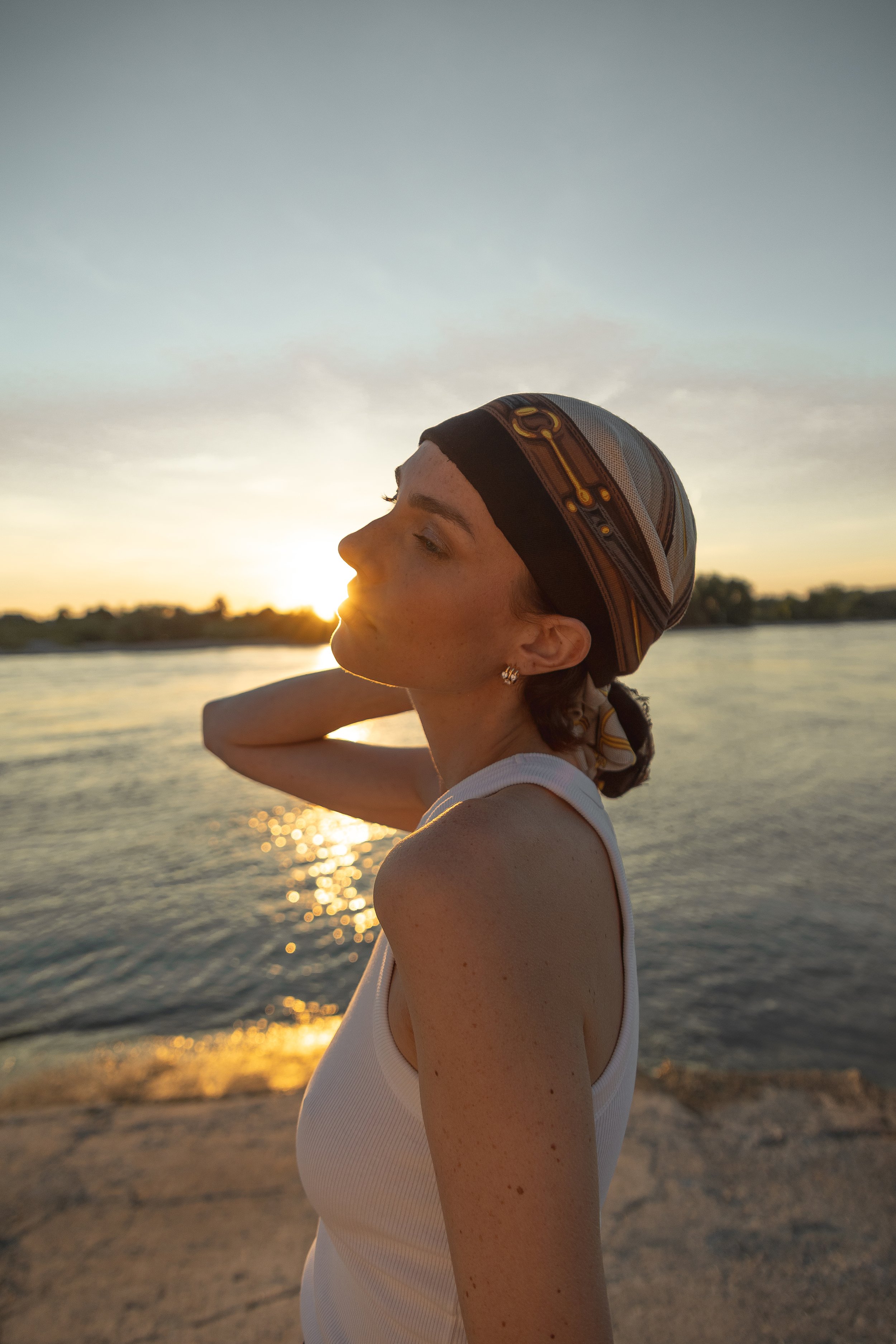 Frau am See bei Sonnenuntergang, trägt ein schwarzes und graues Haarband und weißes Top, schaut verträumt nach links.