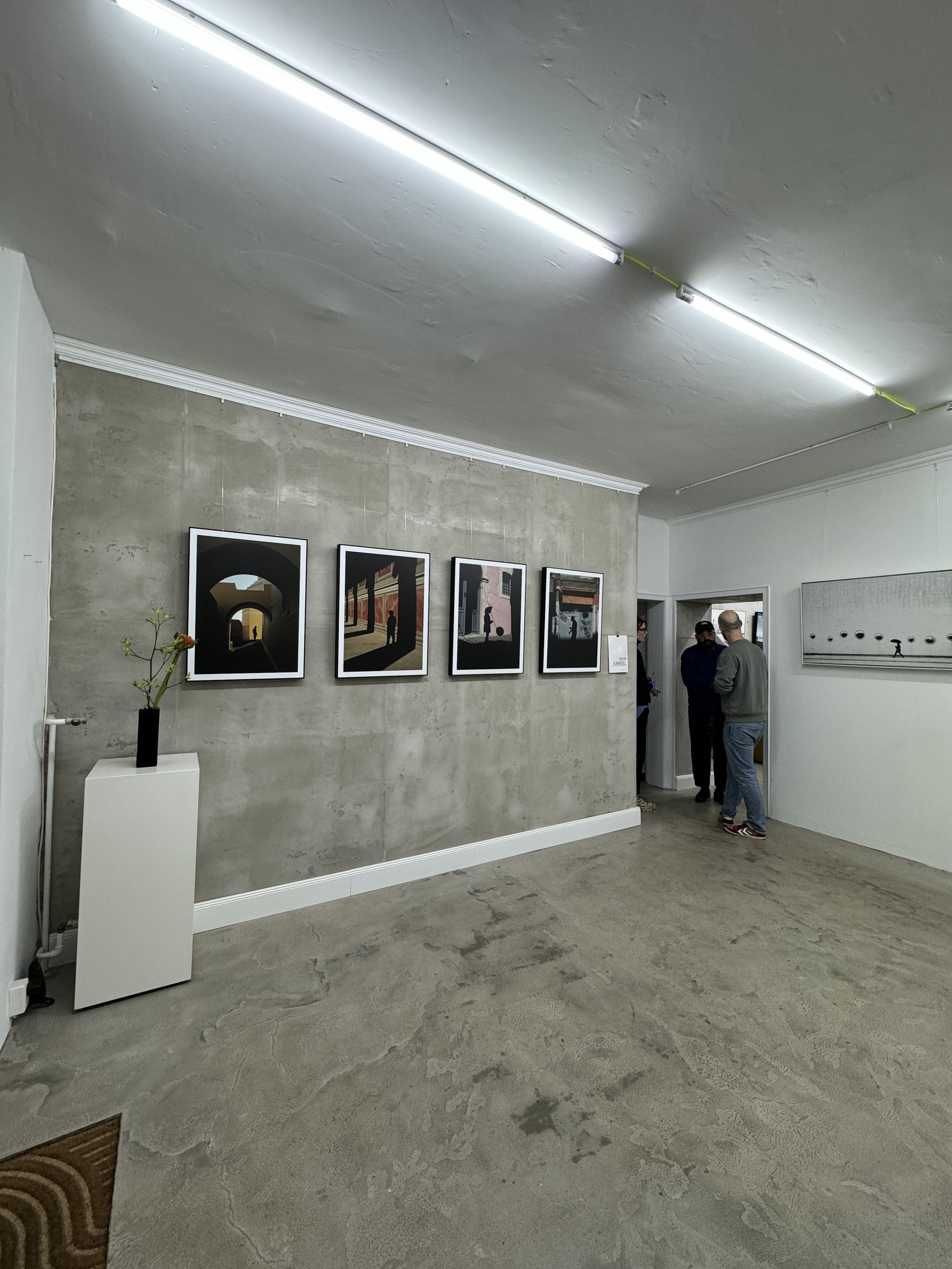 Innenansicht einer modernen Kunstgalerie mit vier Bildern an der Wand, einer weißen Skulptur mit einer Pflanze und mehreren Menschen im Gespräch.