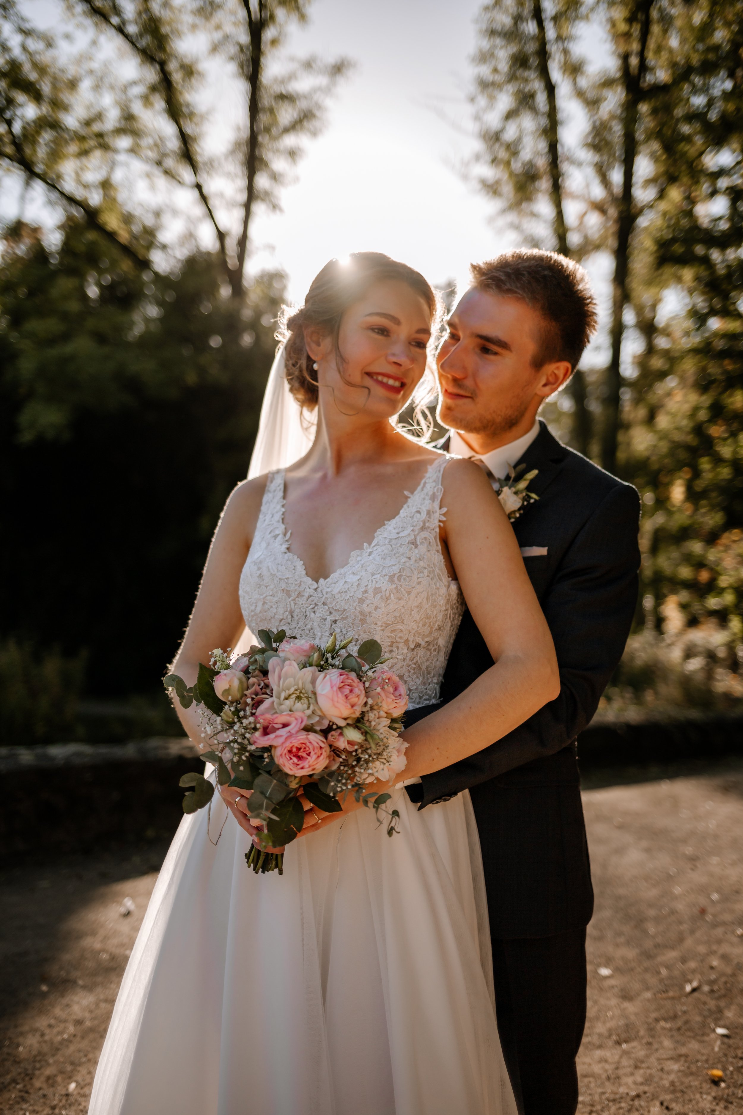 Braut und Bräutigam im Sonnenlicht bei der Hochzeit im Freien, die Braut hält einen Blumenstrauß, umgeben von Bäumen im Herbst.