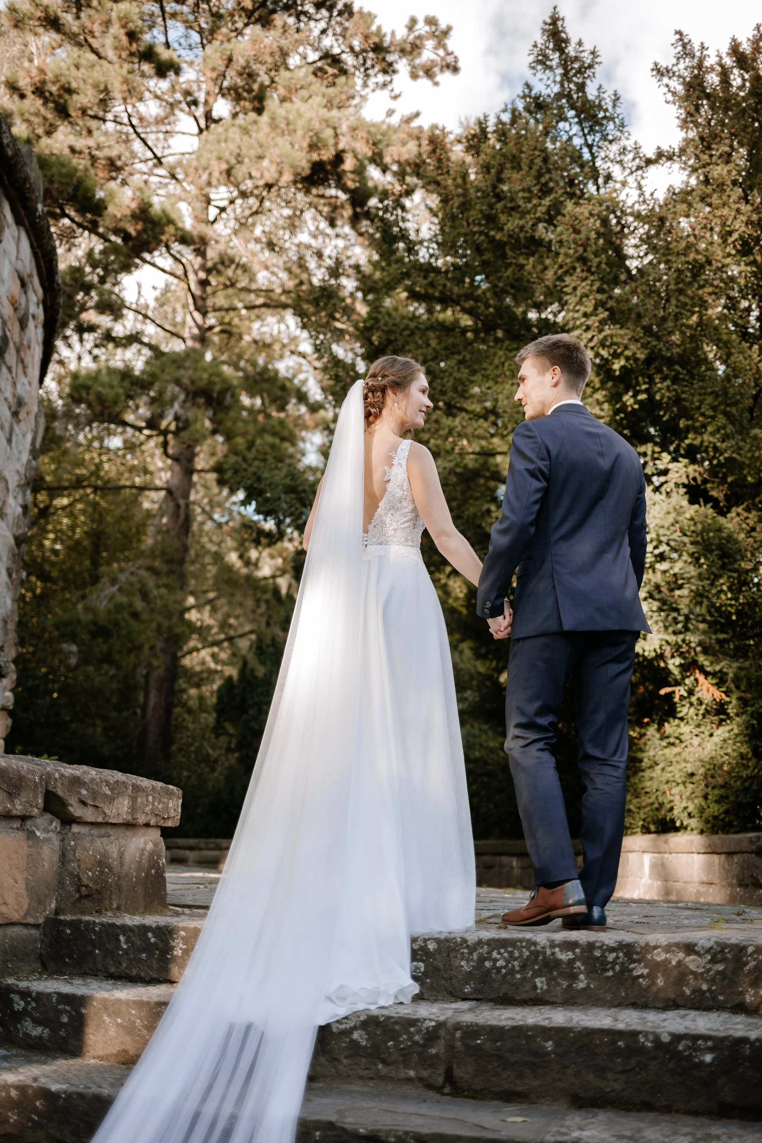 Ein Brautpaar im Freien bei Sonnenuntergang, hält sich an den Händen, die Braut trägt ein langes weißes Hochzeitskleid mit Schleier, der Bräutigam trägt einen dunklen Anzug, umgeben von hohen Bäumen und einem natürlichen Garten.