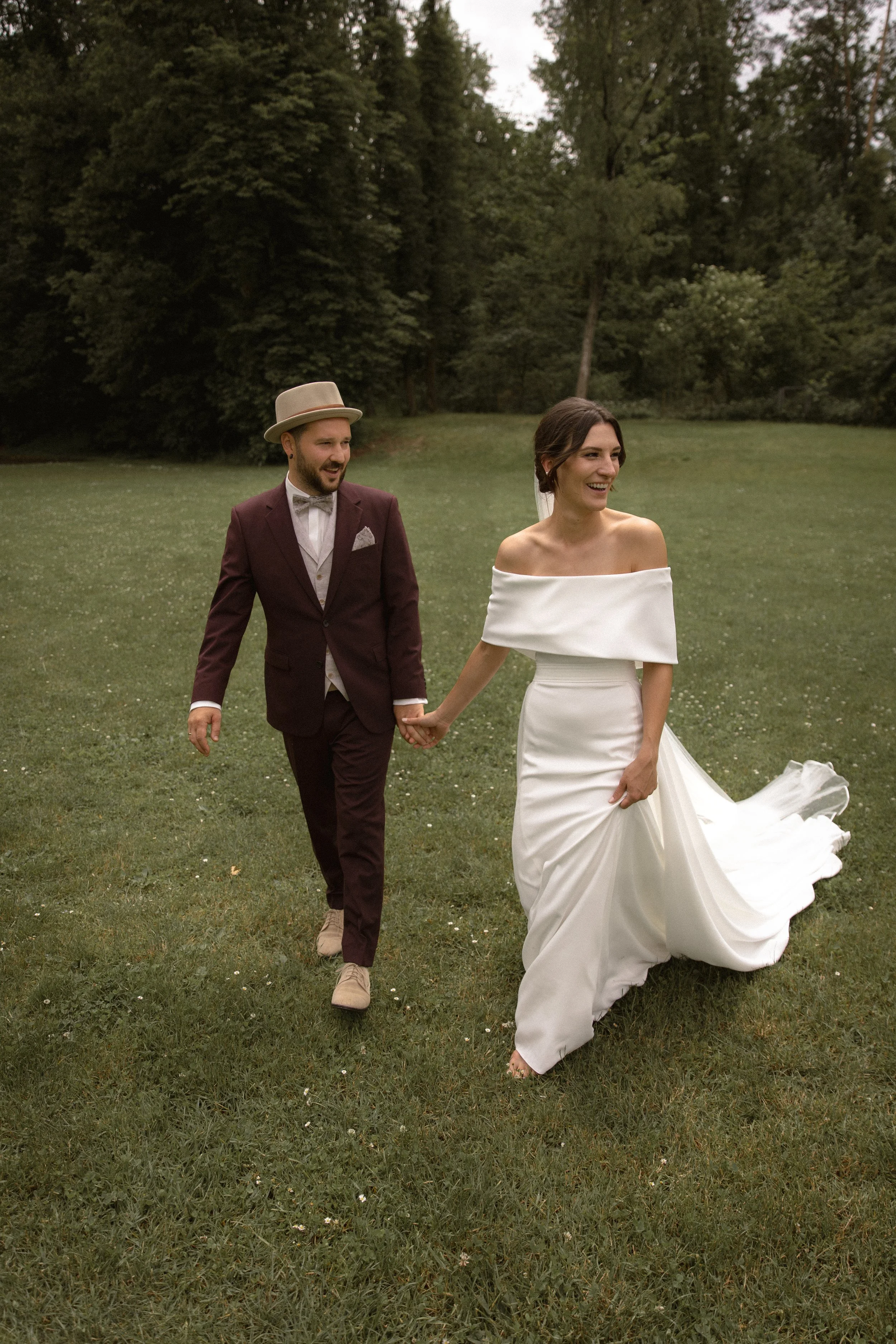 Ein Brautpaar läuft lachend über eine Wiese in einem Park, umgeben von Bäumen, bei ihrer Hochzeit, die Braut trägt ein weißes Hochzeitskleid, der Bräutigam einen Anzug.