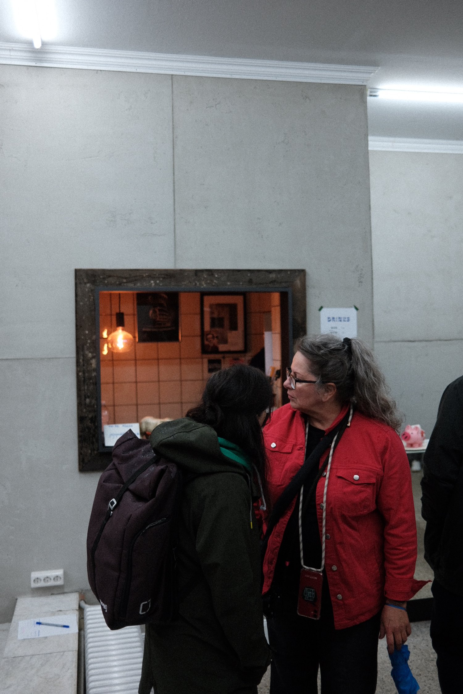 Zwei Frauen sprechen sich in einem Innenraum mit hellen Wänden und einem Wandspiegel an. Eine trägt eine rote Jacke, die andere eine dunkle Jacke mit Rucksack.