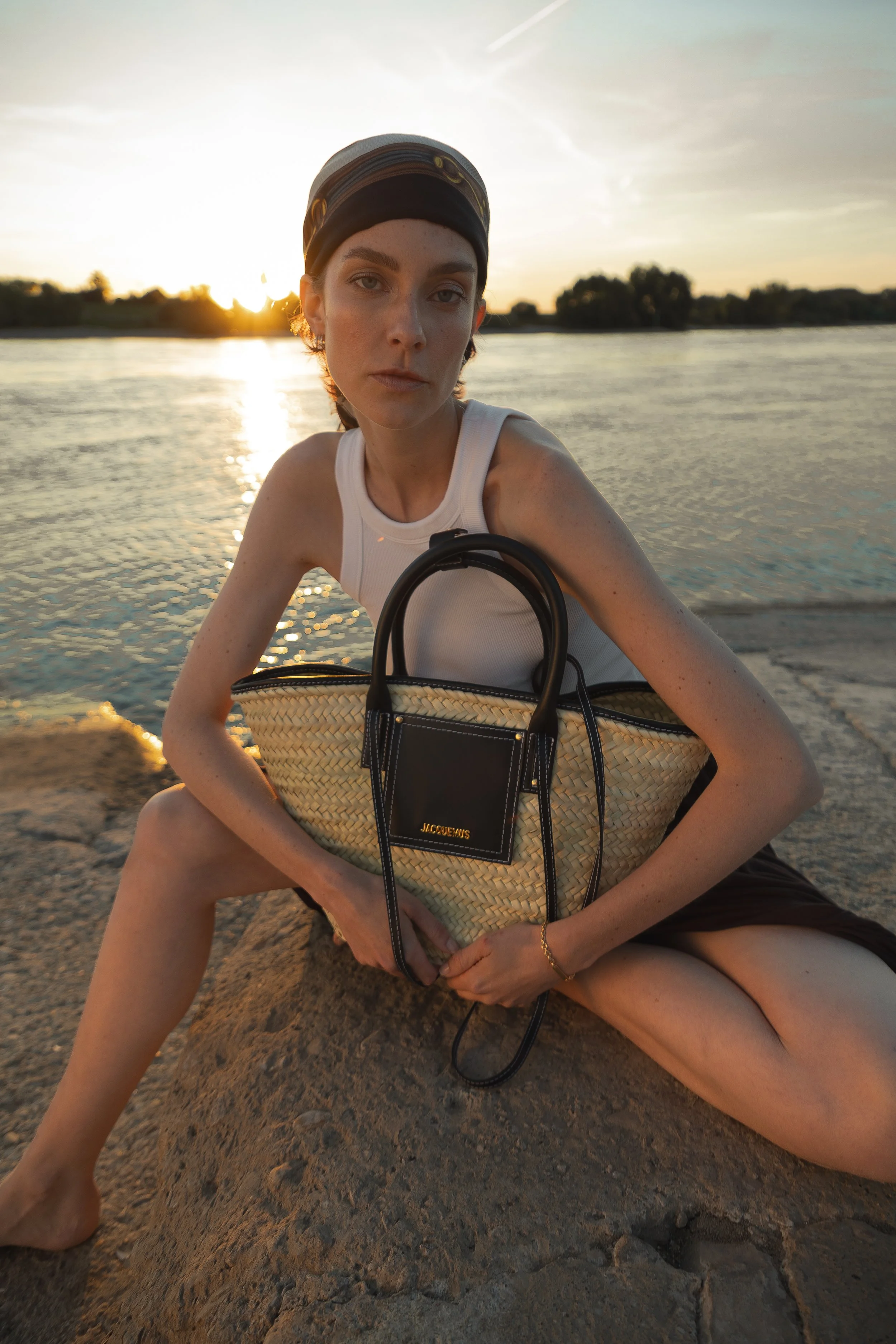 Junge Frau mit Mütze und weißen Top am Flussufer bei Sonnenuntergang, hält eine große Sonnenbrillen- Tasche, sitzt auf einem Felsen.