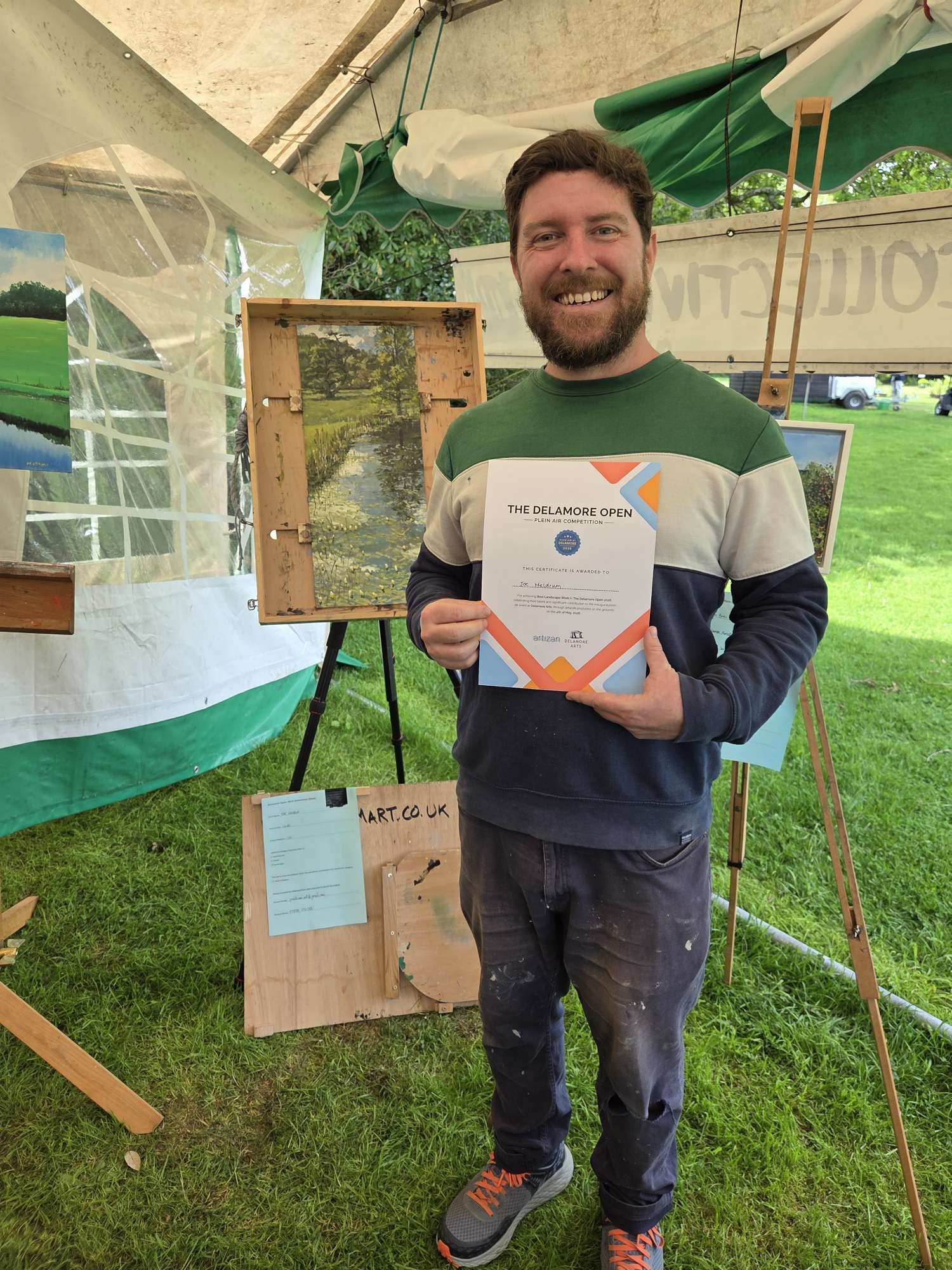 Joe Meldrum Art recieves a certificate for "best landscape" award. He is holding the certificate and standing in front of his painting