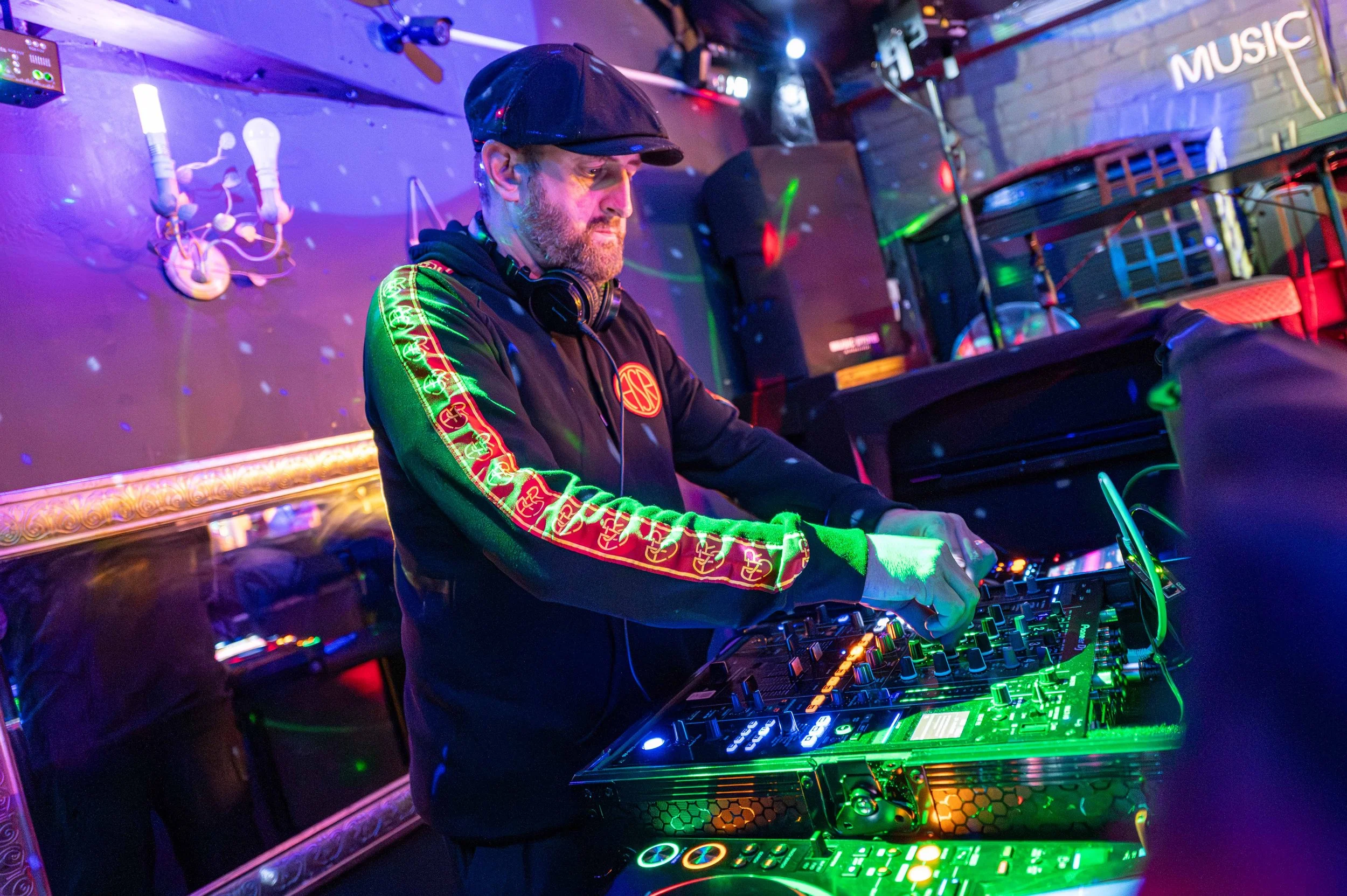 A DJ performing at a nightclub with colorful lighting, wearing headphones and a black hoodie with red and gold designs on the sleeves.