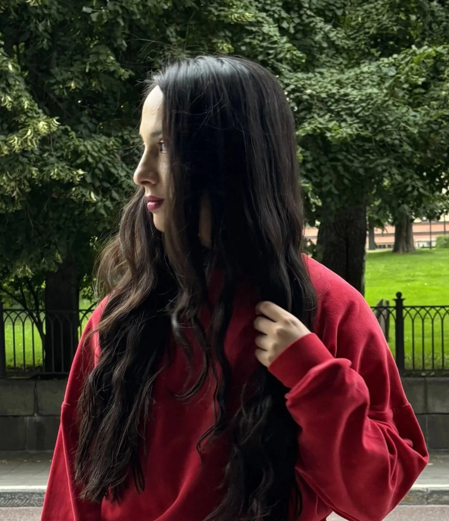 Side profile of a woman with long dark wavy hair wearing a red hoodie, standing outside near a black fence with trees and a green park in the background.
