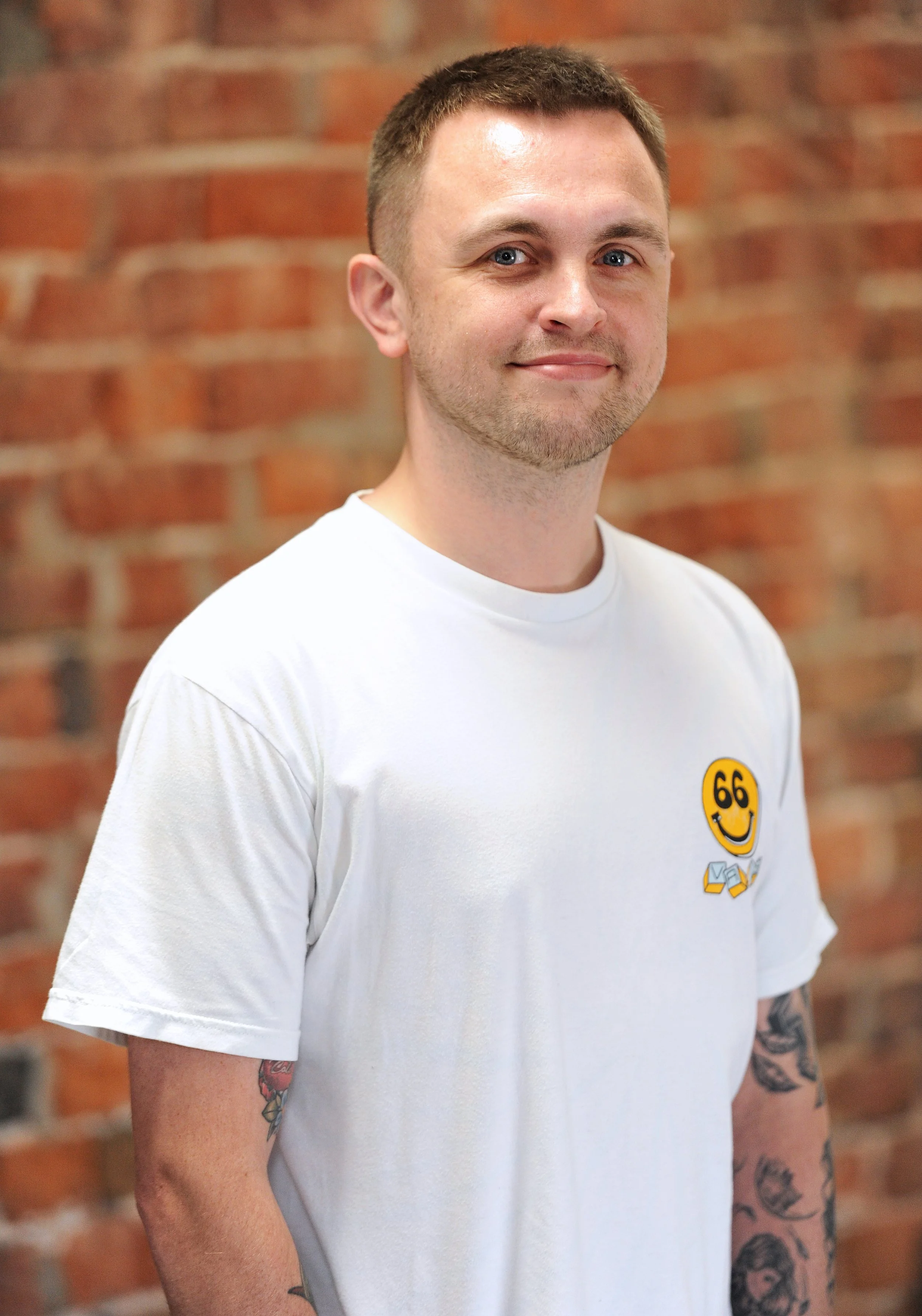 A young man with short brown hair and tattoos on both arms, wearing a white T-shirt with a smiley face and the number 66, standing in front of a brick wall.