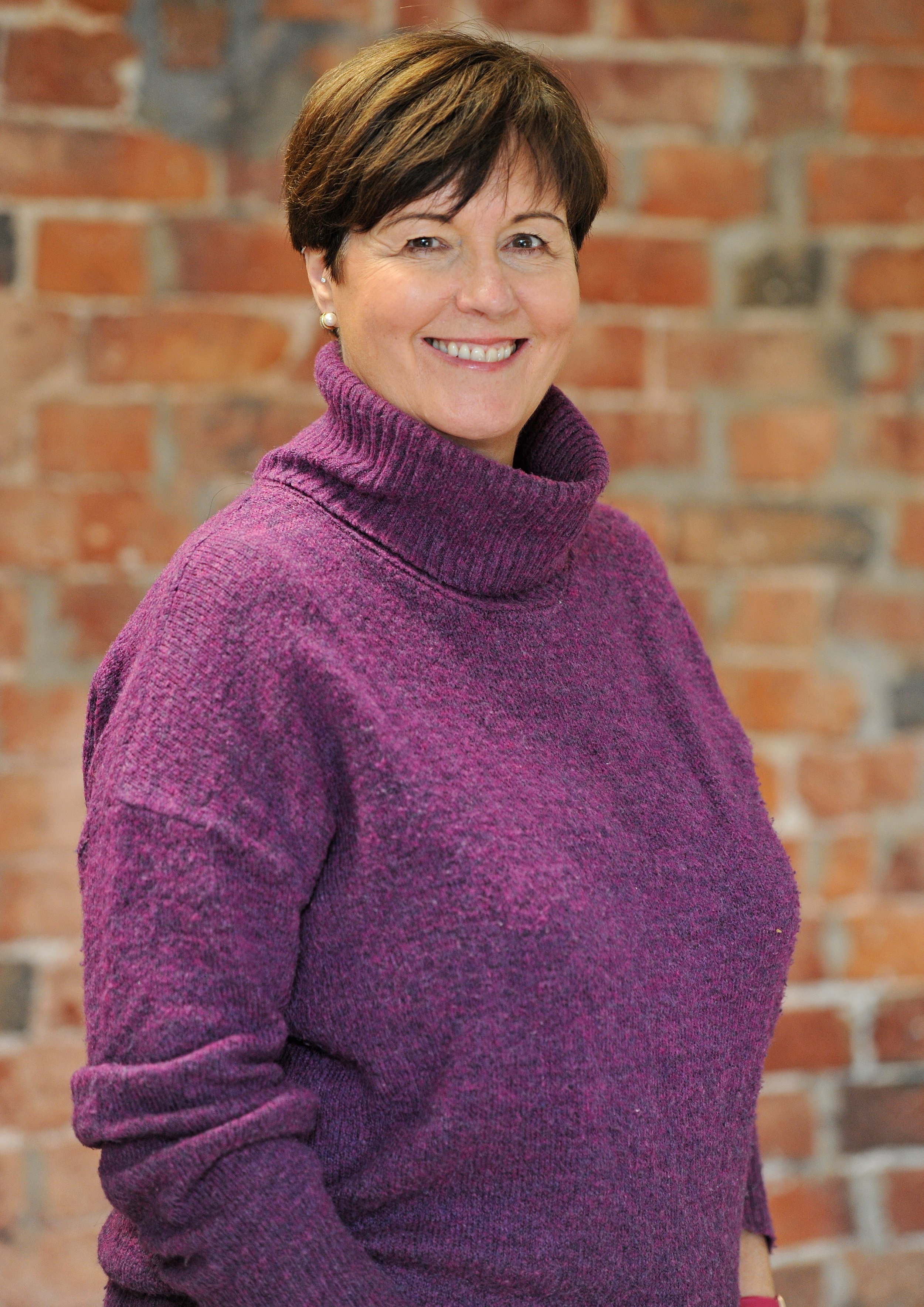 Smiling woman with short brown hair and glasses, wearing a green and white striped shirt, standing in front of a brick wall.