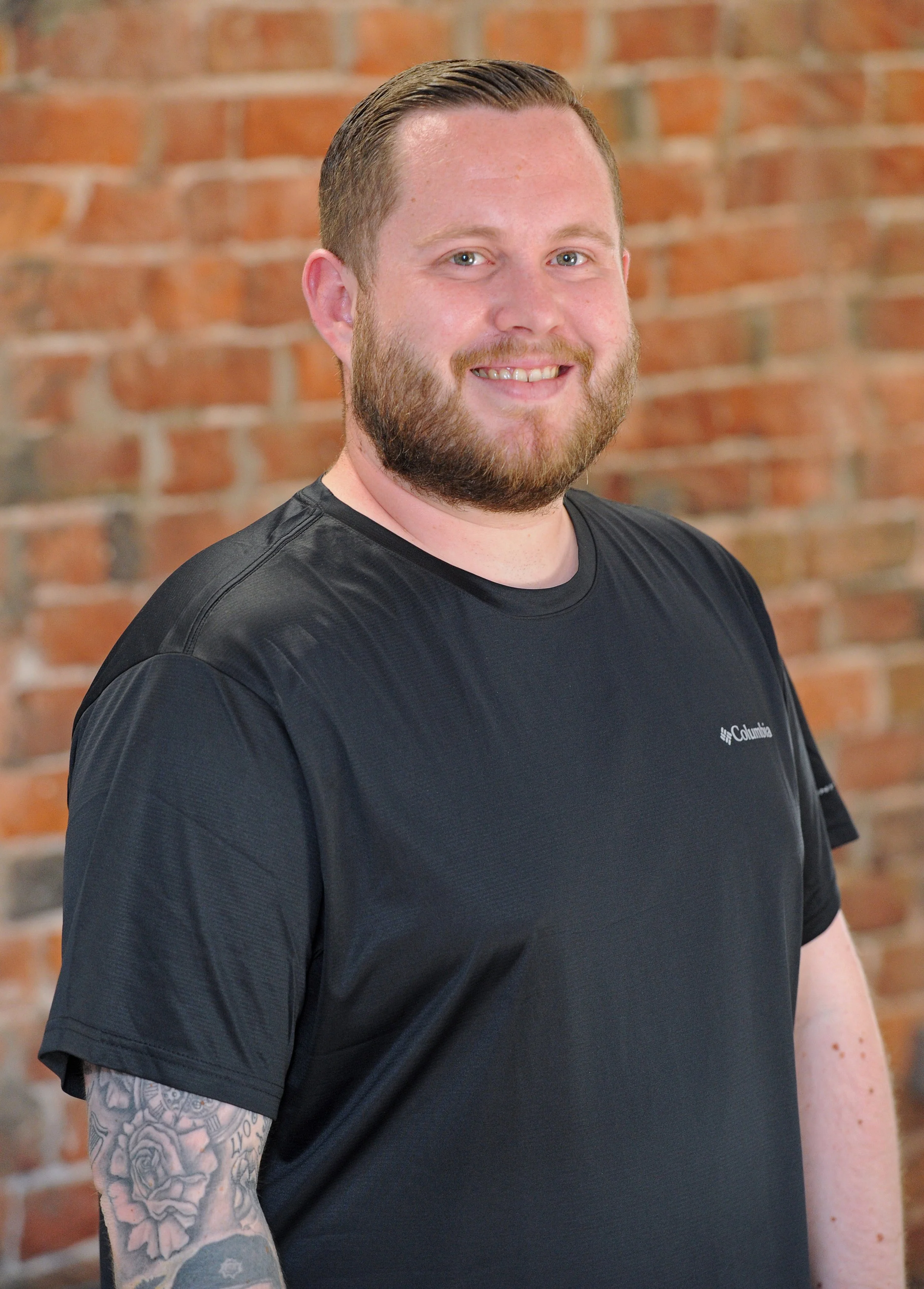 A man with short hair, a beard, and tattoos on his arm, smiling and standing in front of a brick wall, wearing a black Columbia t-shirt.