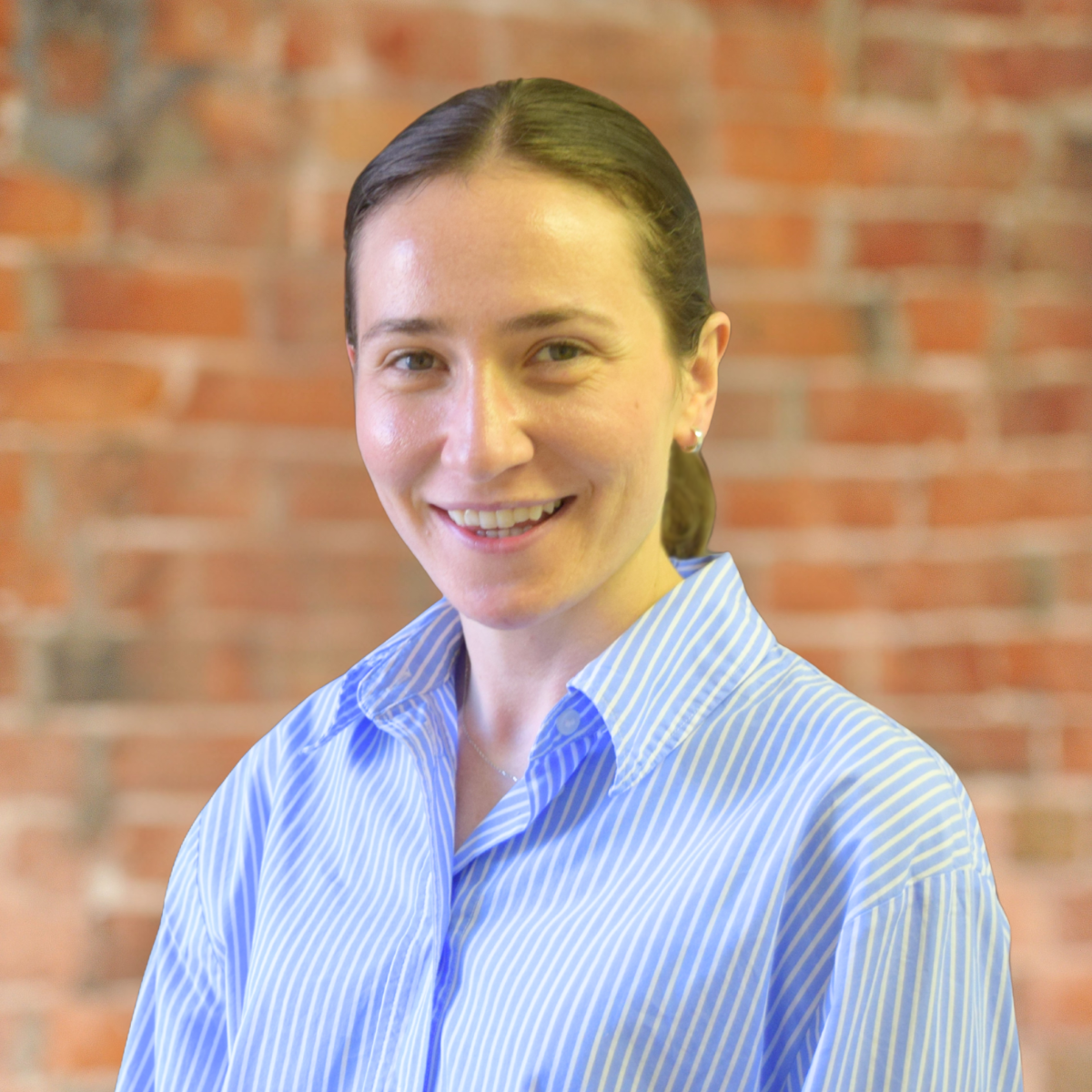 A young woman with long brown hair, wearing a dark headband and maroon sweater, smiling at the camera against a light-colored brick wall.