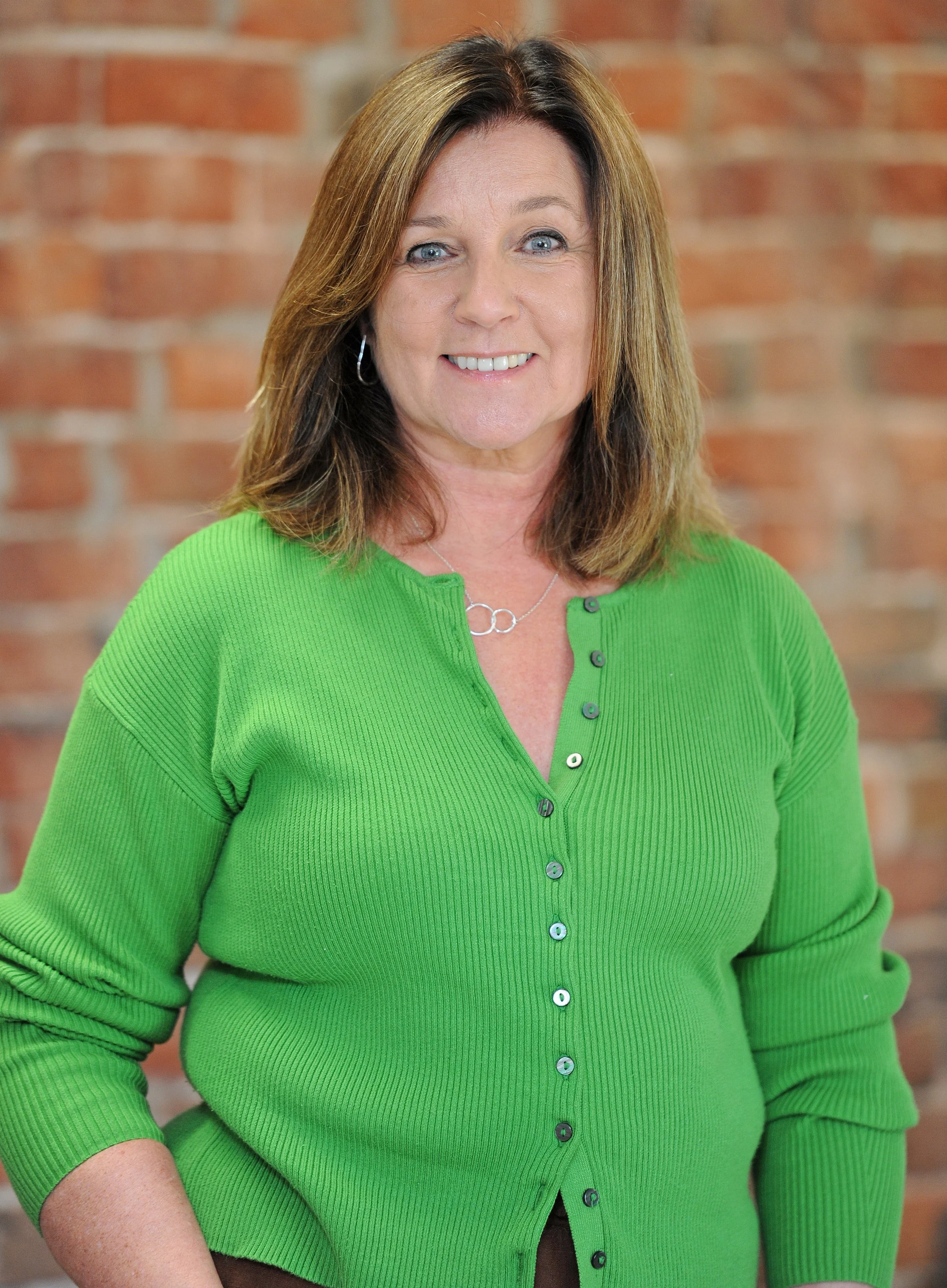 A woman with shoulder-length brown hair, blue eyes, and fair skin, wearing a bright green ribbed button-up sweater, and smiling, standing in front of a brick wall.