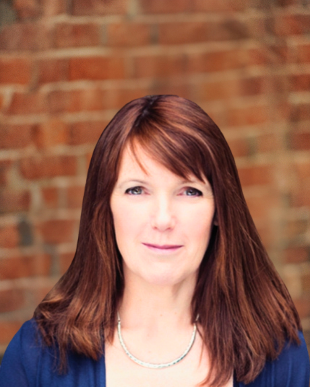A smiling middle-aged woman with short blonde hair and blue eyes, wearing a patterned short-sleeve top, standing against a brick wall background.