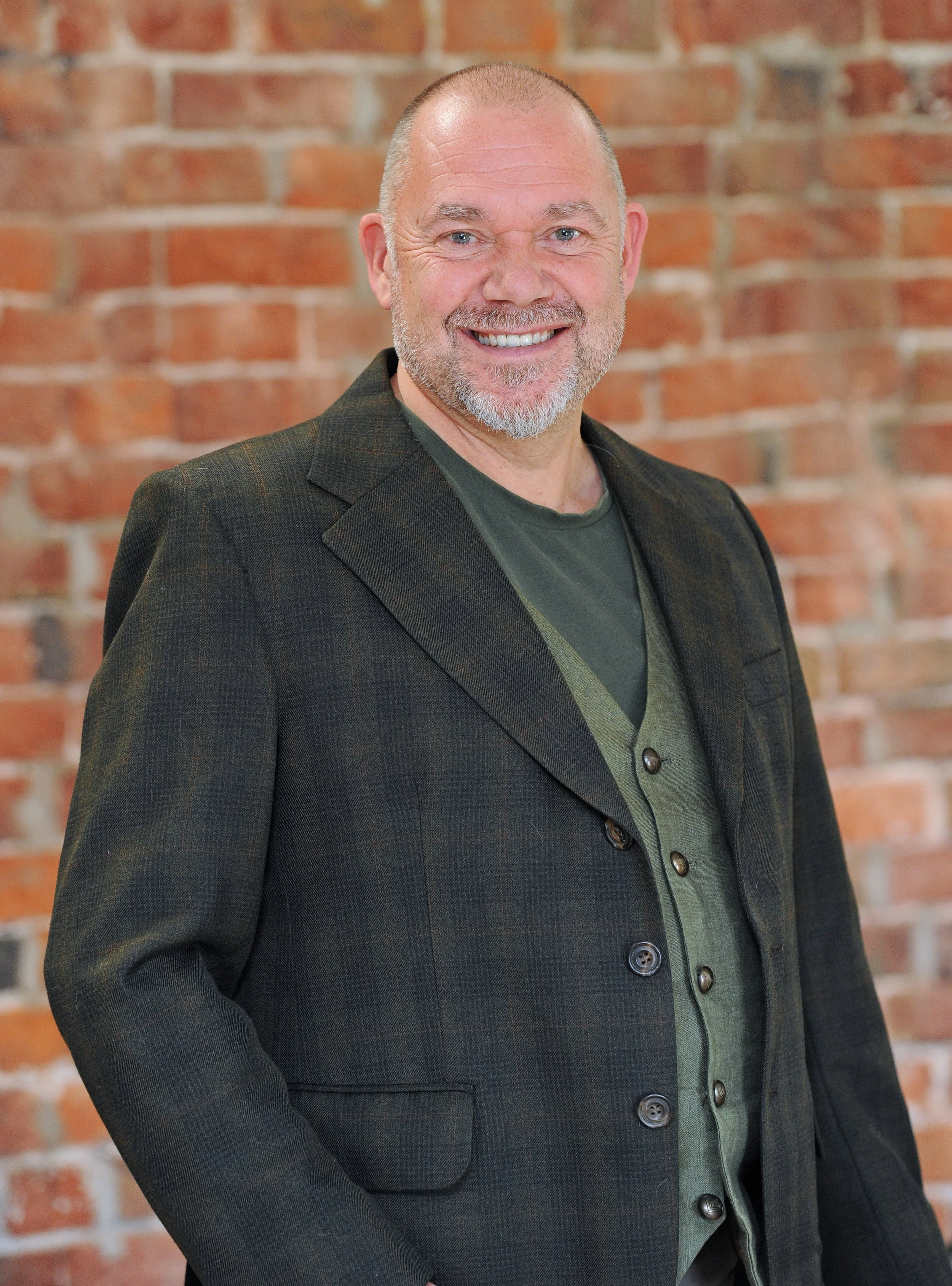 A middle-aged man with a smile, wearing a blue vest over a collared shirt, standing against a light-colored brick wall.