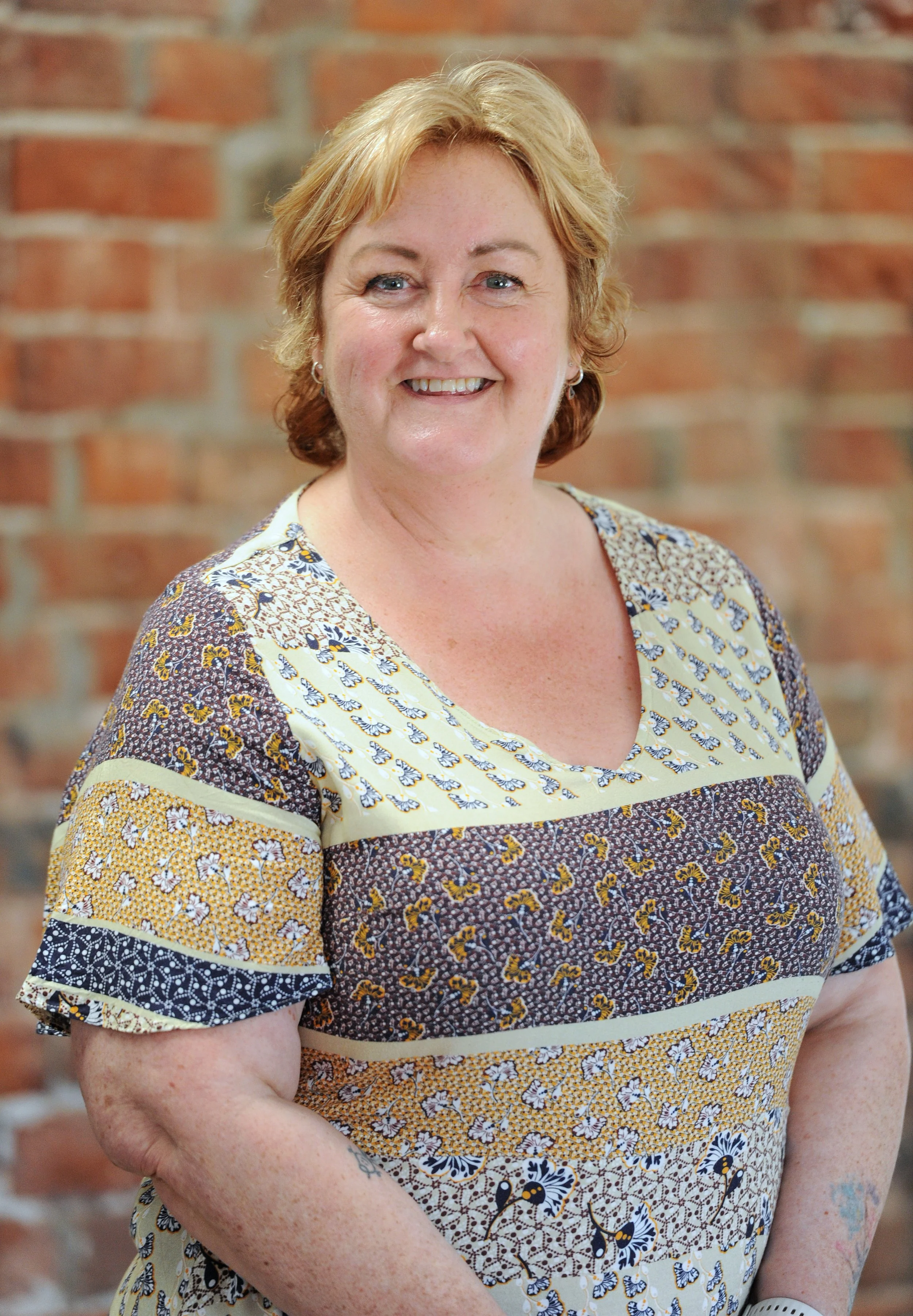 A smiling middle-aged woman with short blonde hair and blue eyes, wearing a patterned short-sleeve top, standing against a brick wall background.