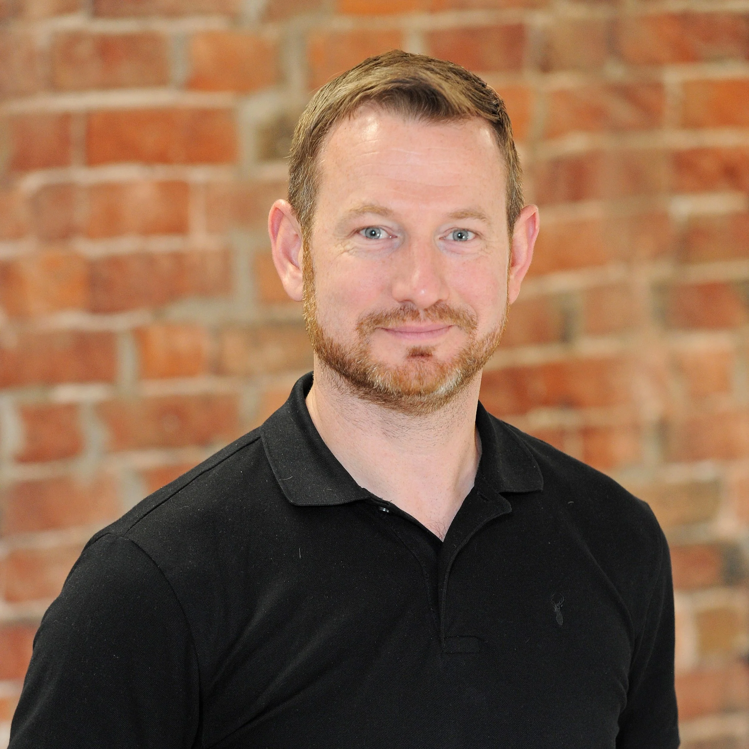 A man with short brown hair and a beard, wearing a black polo shirt, standing in front of a brick wall.