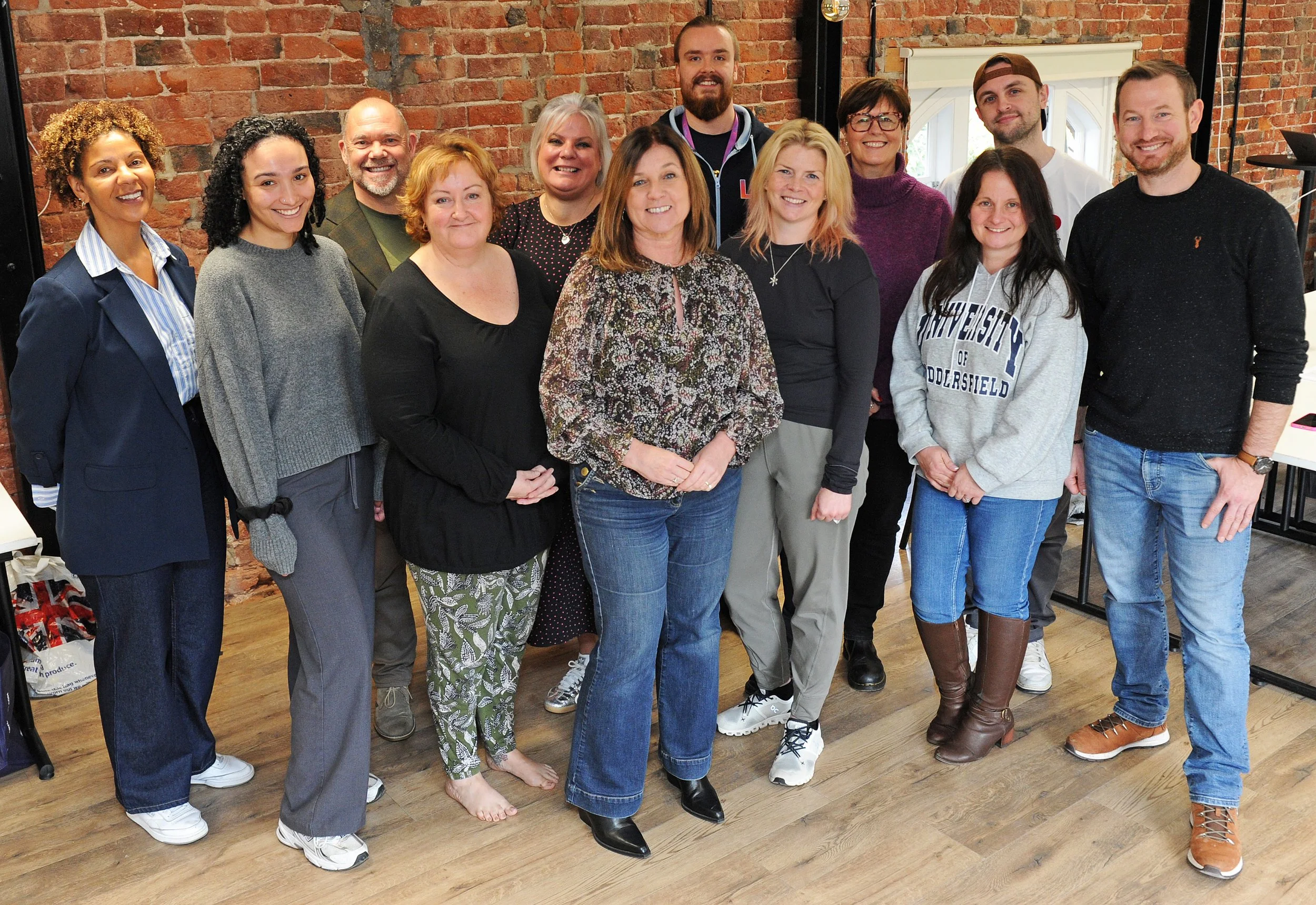 Group of fifteen people standing together indoors in front of a brick wall, smiling at the camera, with some wearing casual clothing and sneakers, and others in more formal attire.