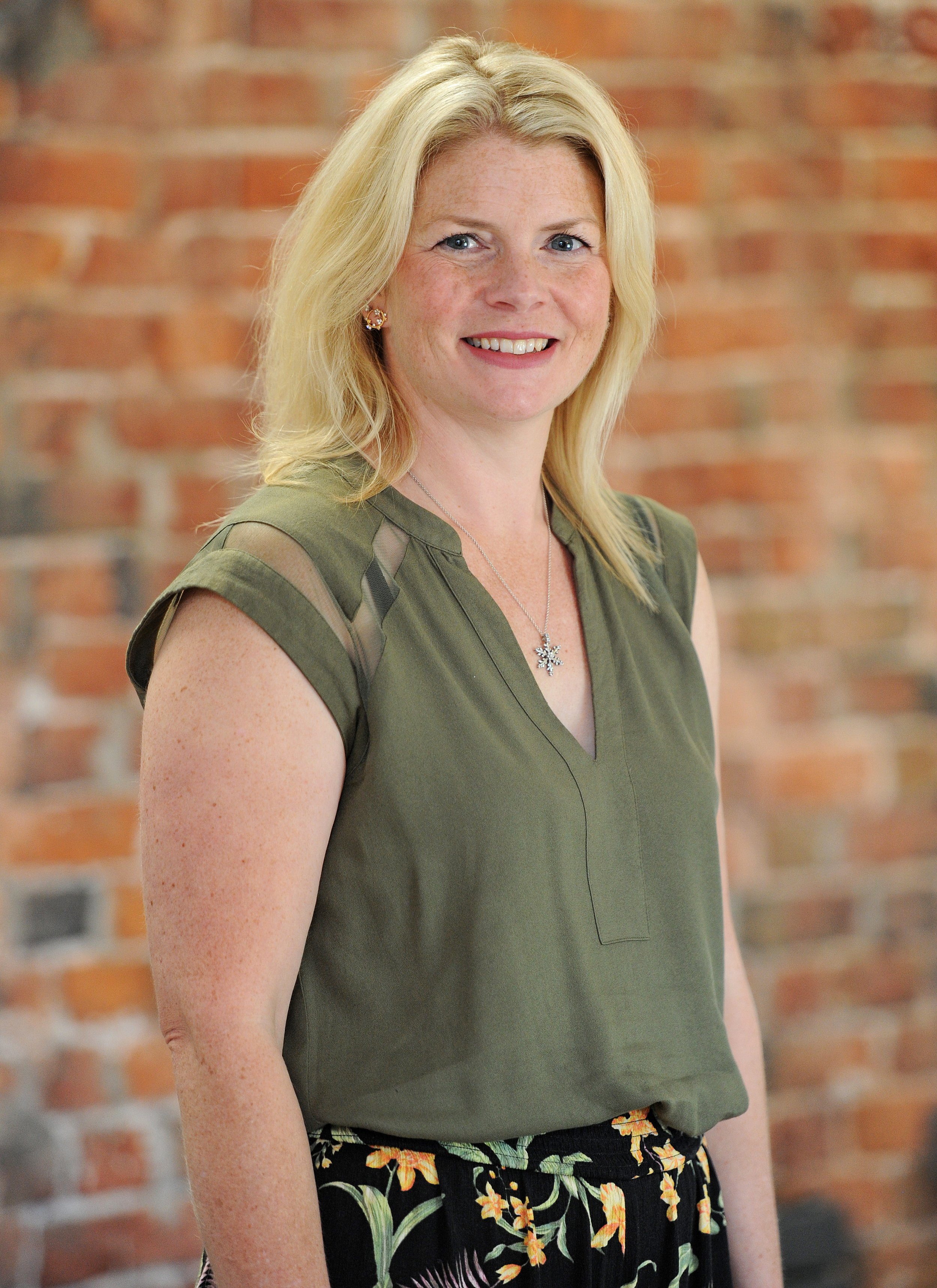 A smiling woman with blonde hair, wearing a green blouse with cut-out shoulder details, a necklace with a snowflake pendant, and floral patterned pants, standing in front of a brick wall.