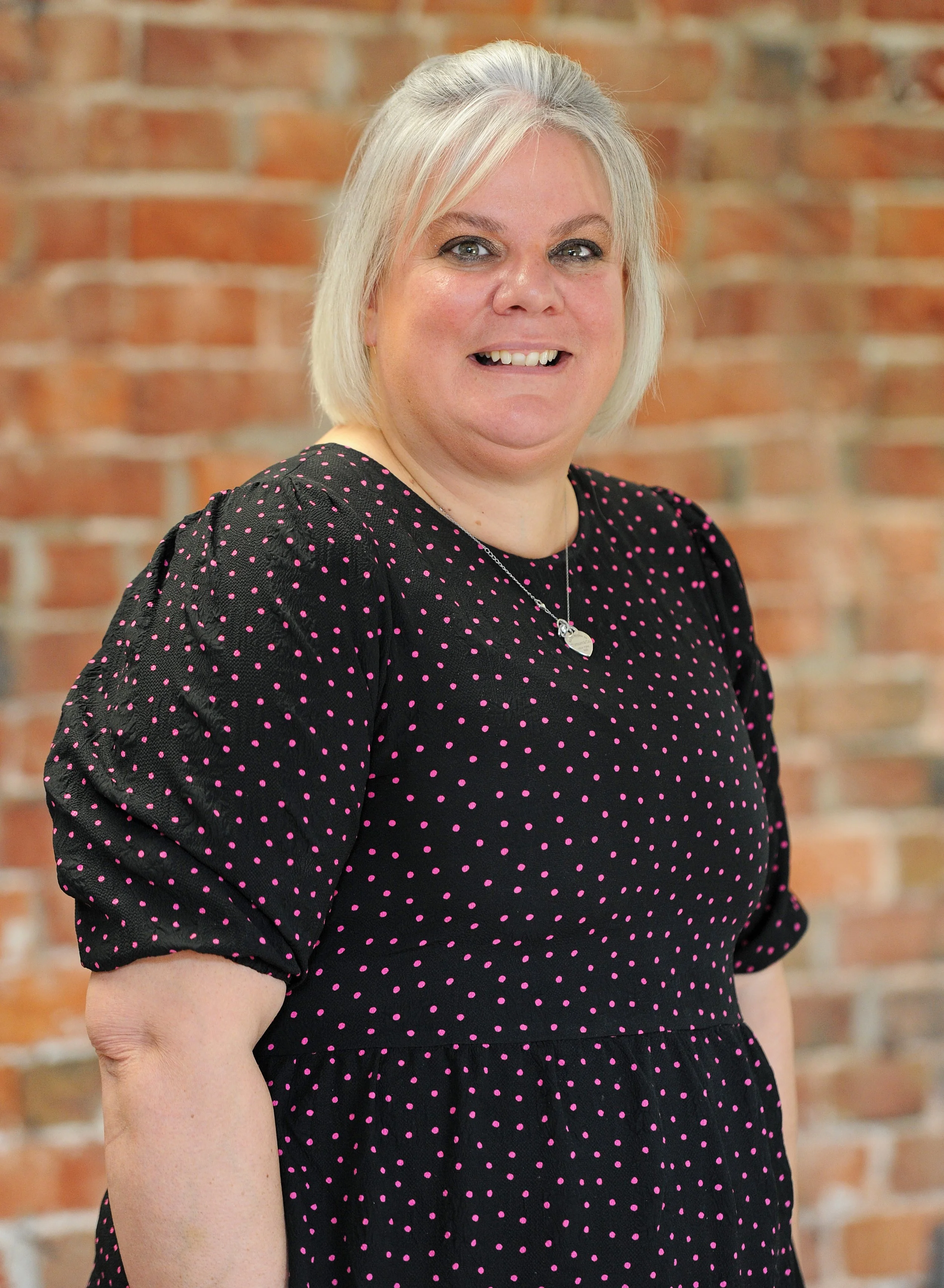 A woman with blonde hair smiling, wearing a purple and white patterned dress, standing in front of a brick wall.