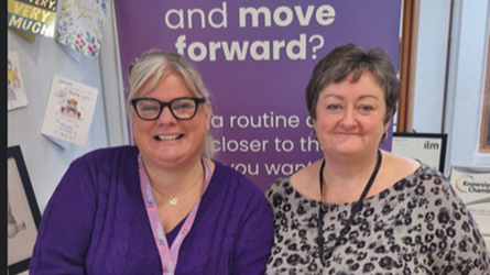 Two smiling women standing in front of a purple sign that says 'and move forward?' with additional text that is partially visible.