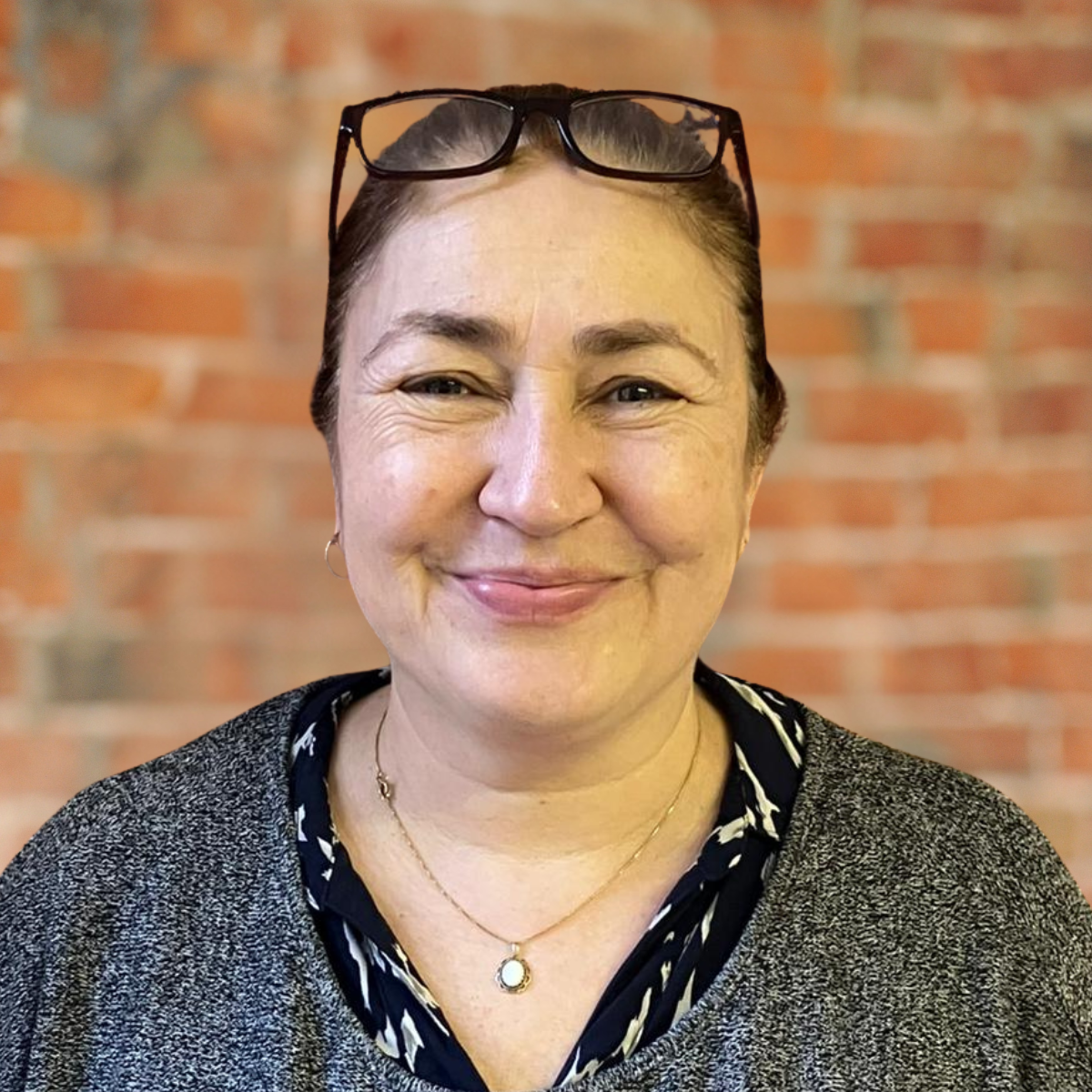 A woman smiling in front of a brick wall. She is wearing glasses on her head, a gray sweater, a patterned shirt, and a gold necklace.