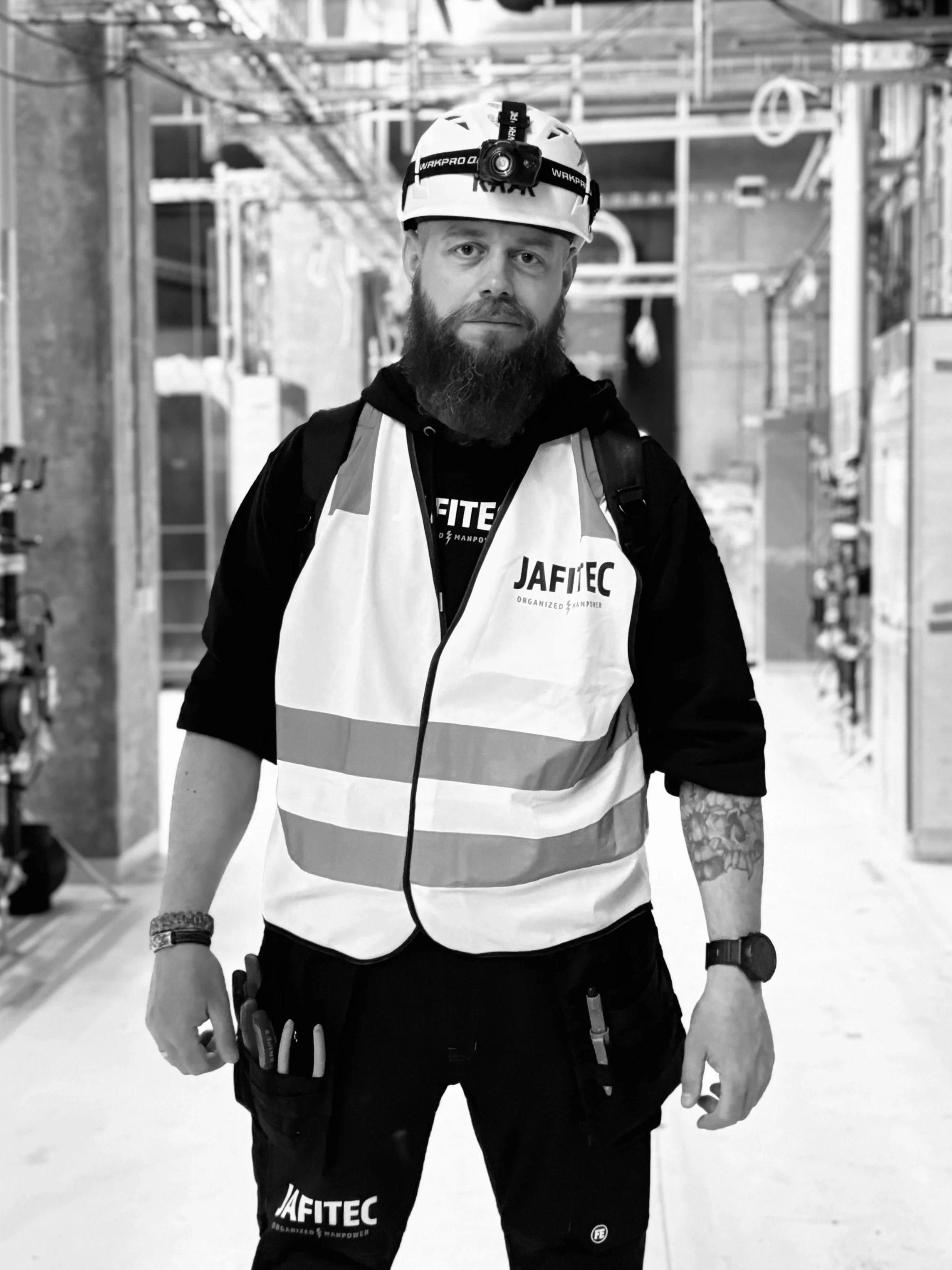 A construction worker with a beard and tattoos wearing a helmet with a headlamp, a reflective safety vest, and black work pants standing inside a building under construction.