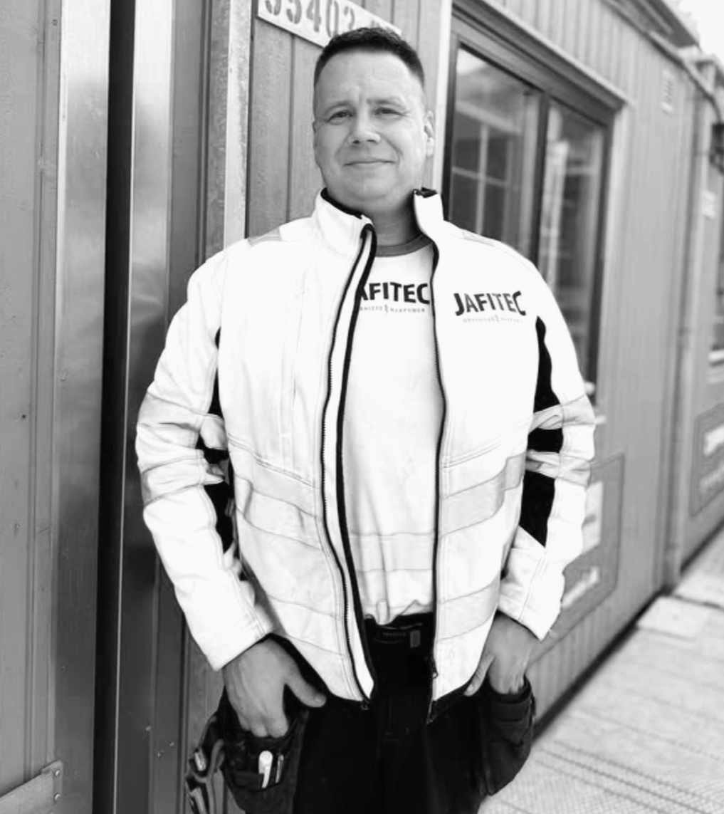 A smiling construction worker wearing a safety helmet and a high-visibility jacket, standing inside an industrial or construction site.